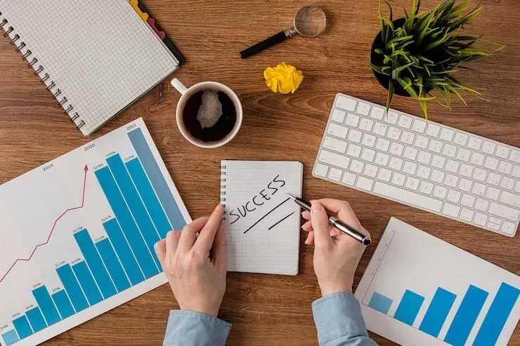 Hands analyzing growth charts at a desk with laptop, notebook, coffee, and plant