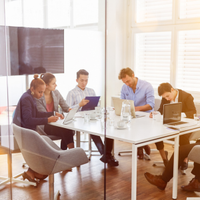 Five coworkers seated around a white table in a bright office meeting room, reviewing laptops and papers.