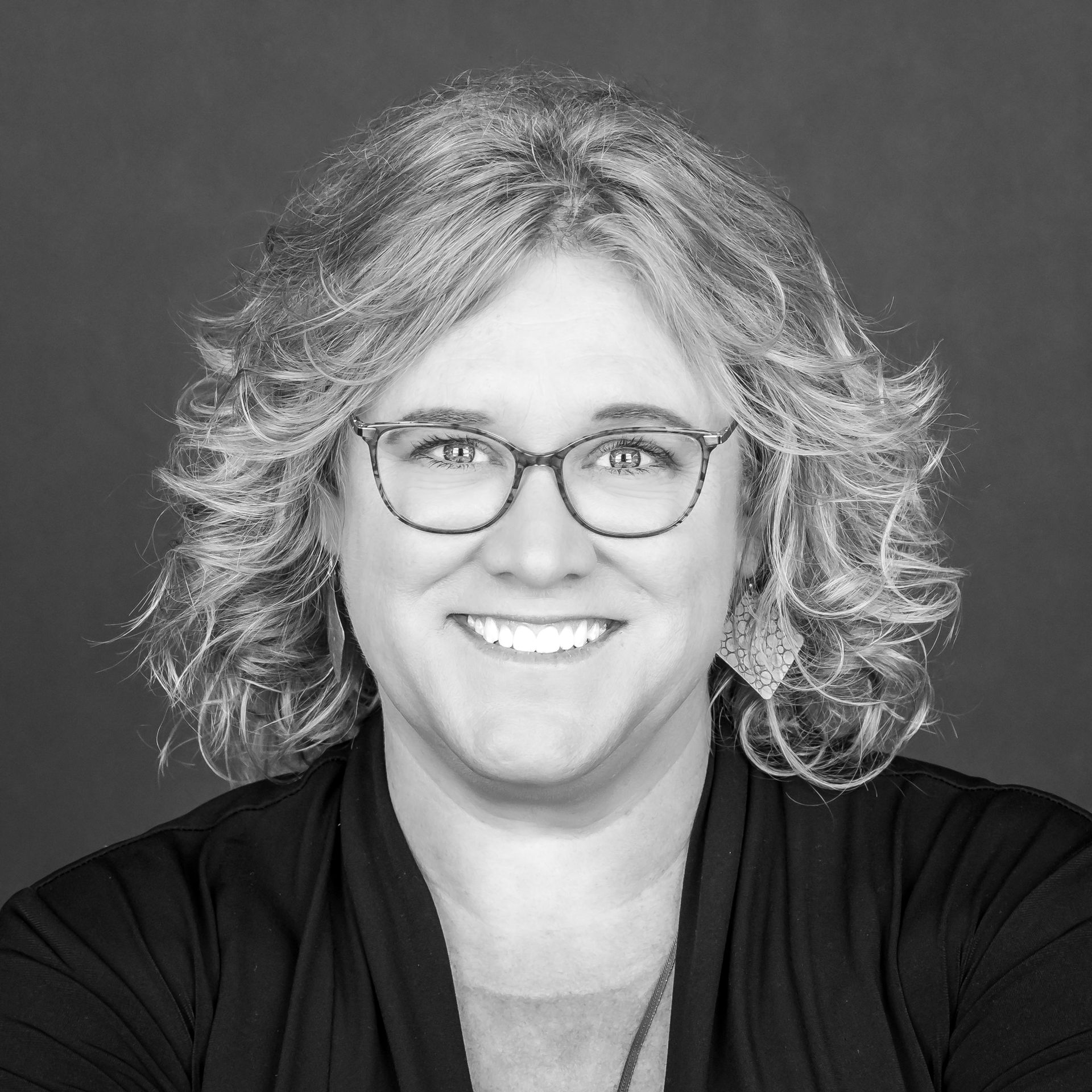 Smiling person with glasses and curly hair in a black top on a gray background