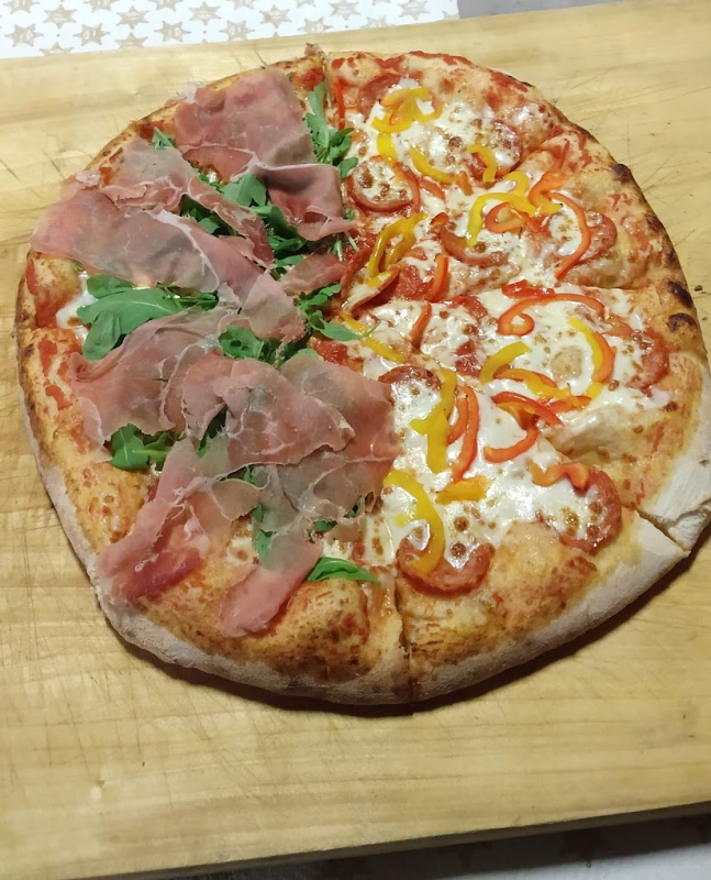 Pizza metà e metà con prosciutto e rucola da un lato, peperoni e formaggio fuso dall'altro, su un tagliere di legno.