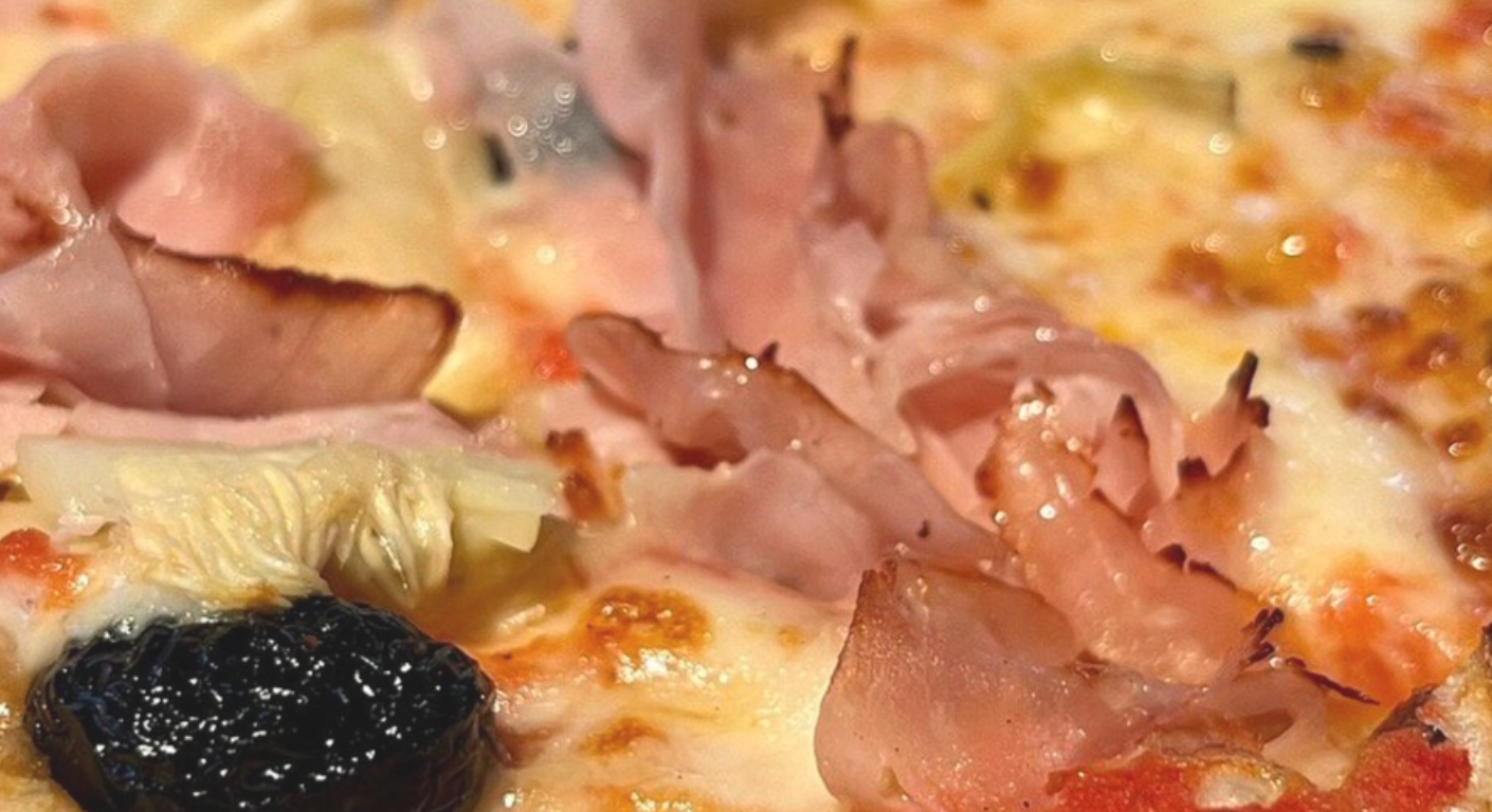 Primo piano di una pizza condita con formaggio fuso, pezzi di prosciutto e un'oliva nera.