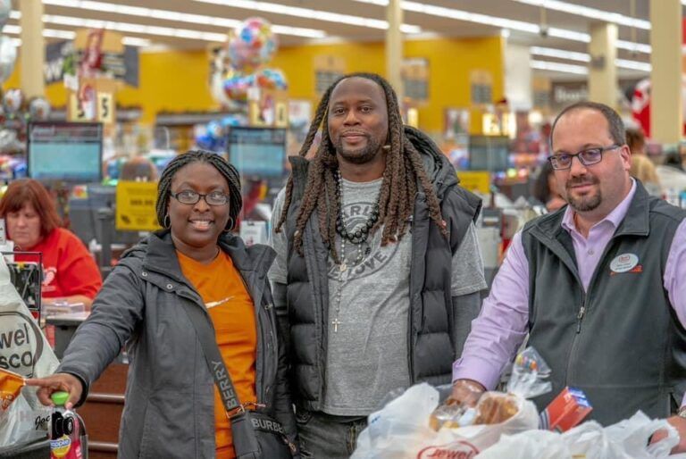 Three people standing with bags of groceries inside a supermarket, smiling at the camera.