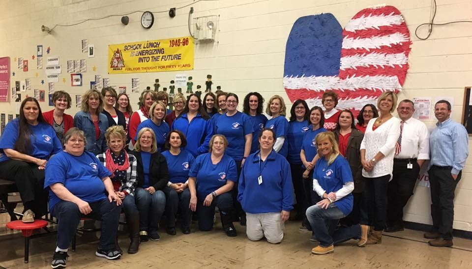 Group of adults posing in a classroom, many in blue shirts, with an American flag mural on the wall.