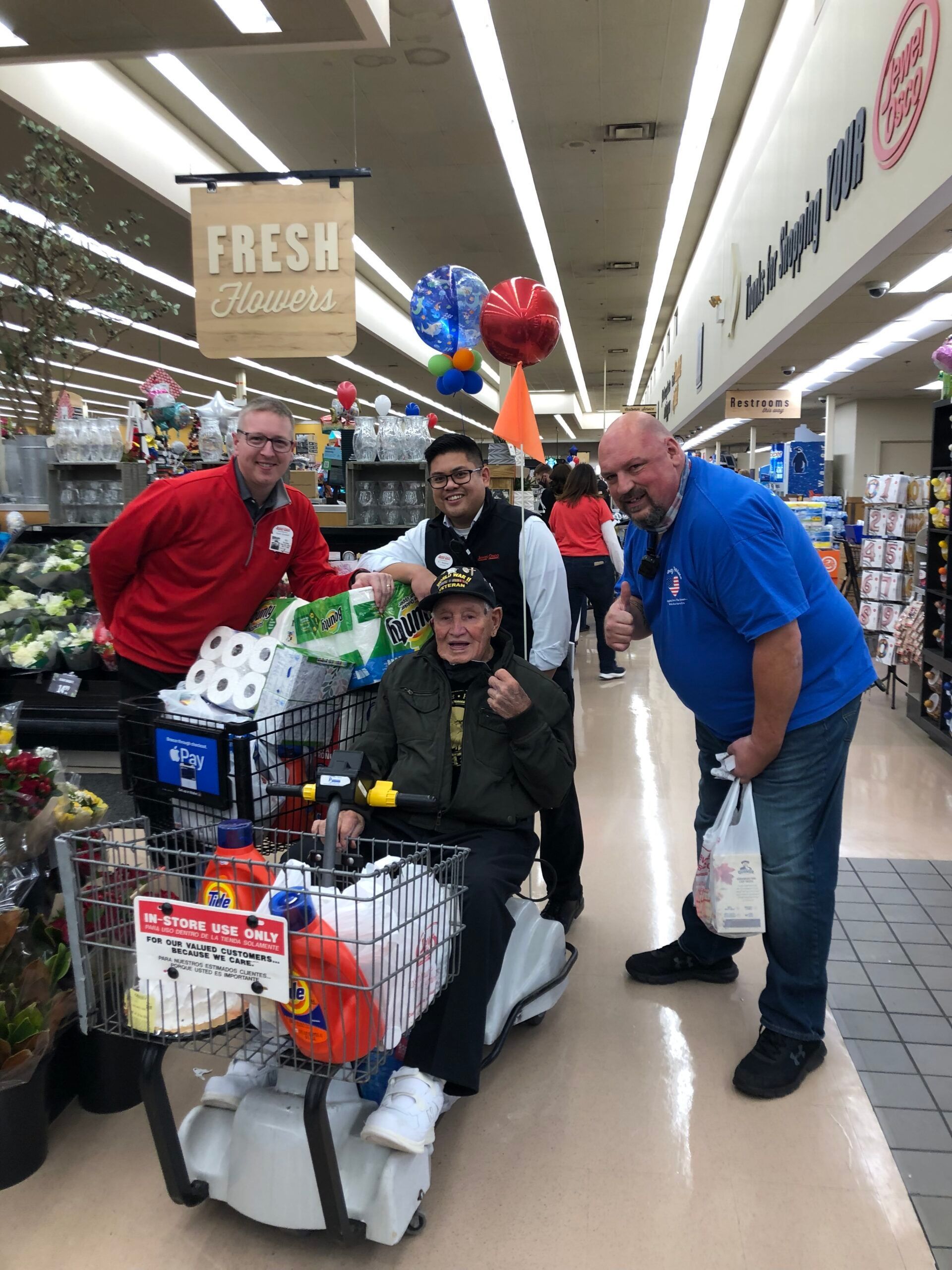 People in a grocery store aisle with a decorated shopping cart, balloons, and a large “Fresh” sign.