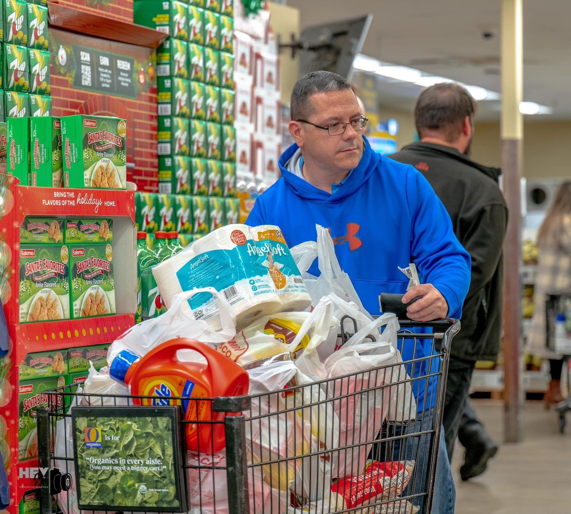 Man in blue hoodie pushing a cart full of groceries in a supermarket aisle