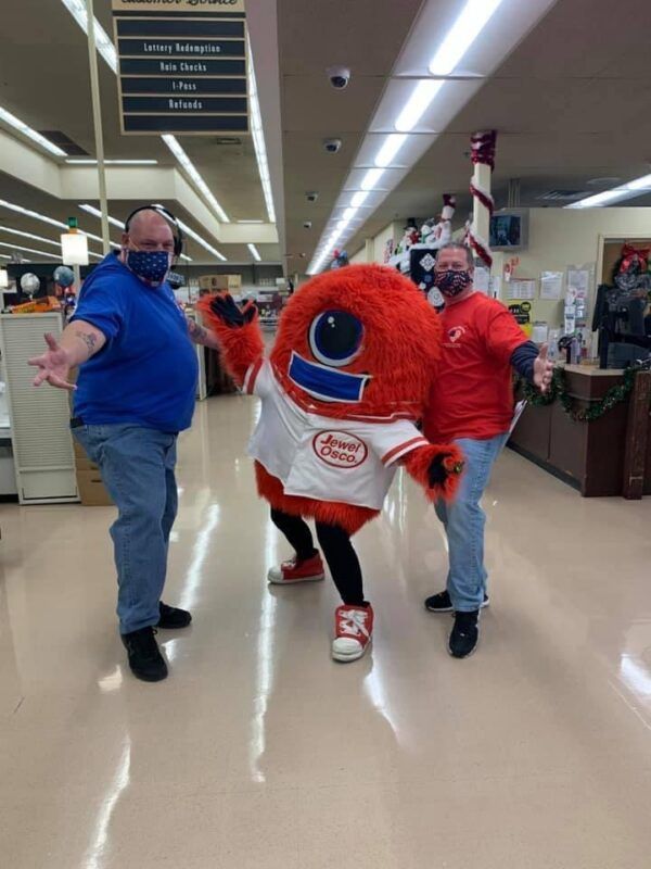 Two masked shoppers in a grocery store with a large red-and-white team mascot between them