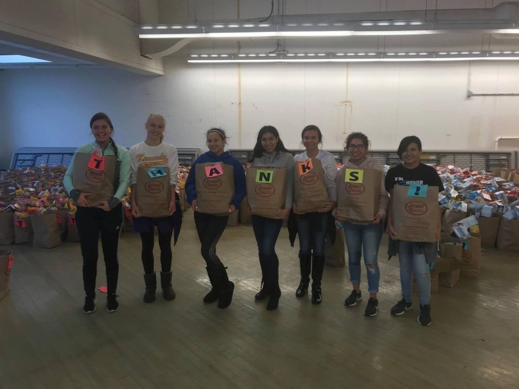 Seven volunteers in numbered aprons stand in a warehouse in front of rows of bagged donations.