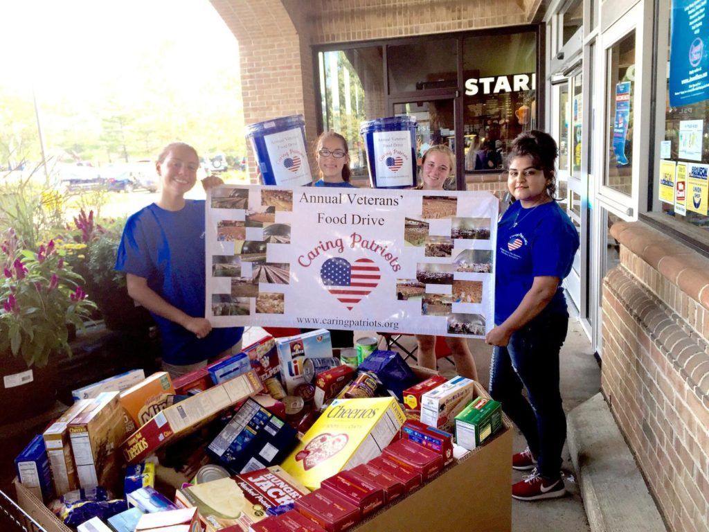 Two volunteers hold a food drive sign beside a table piled with donated groceries.