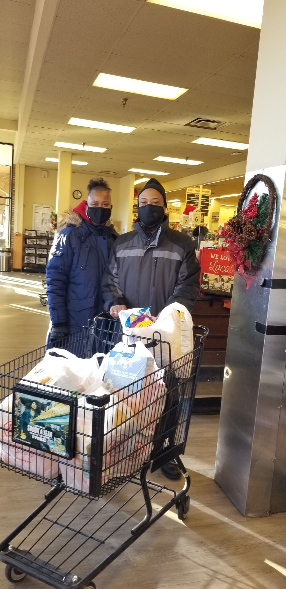 Two masked people stand behind a shopping cart full of bags in a store entrance.