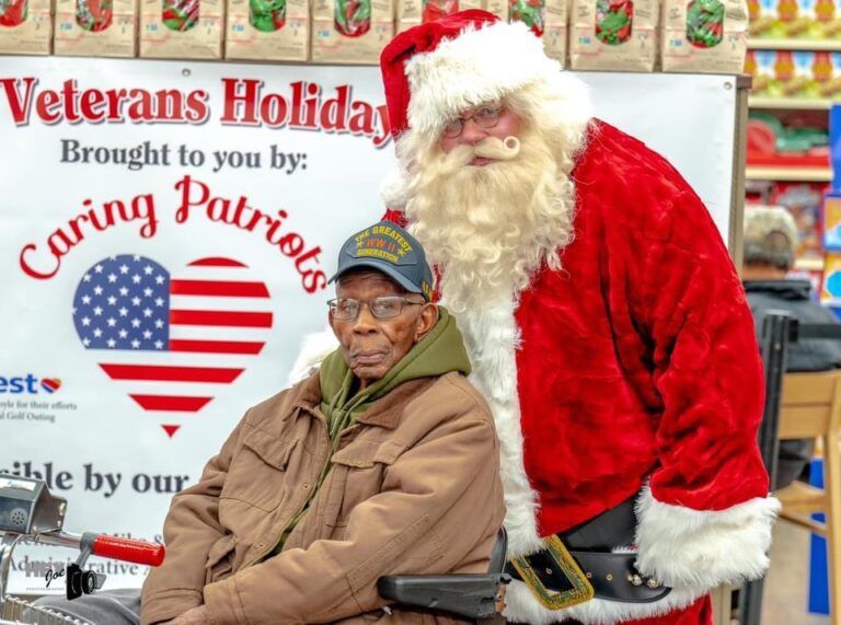 Veterans holiday event with Santa standing behind a seated man in front of a Caring Patriots banner