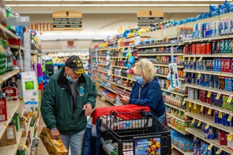 Two shoppers talking in a grocery store aisle beside a cart full of red items