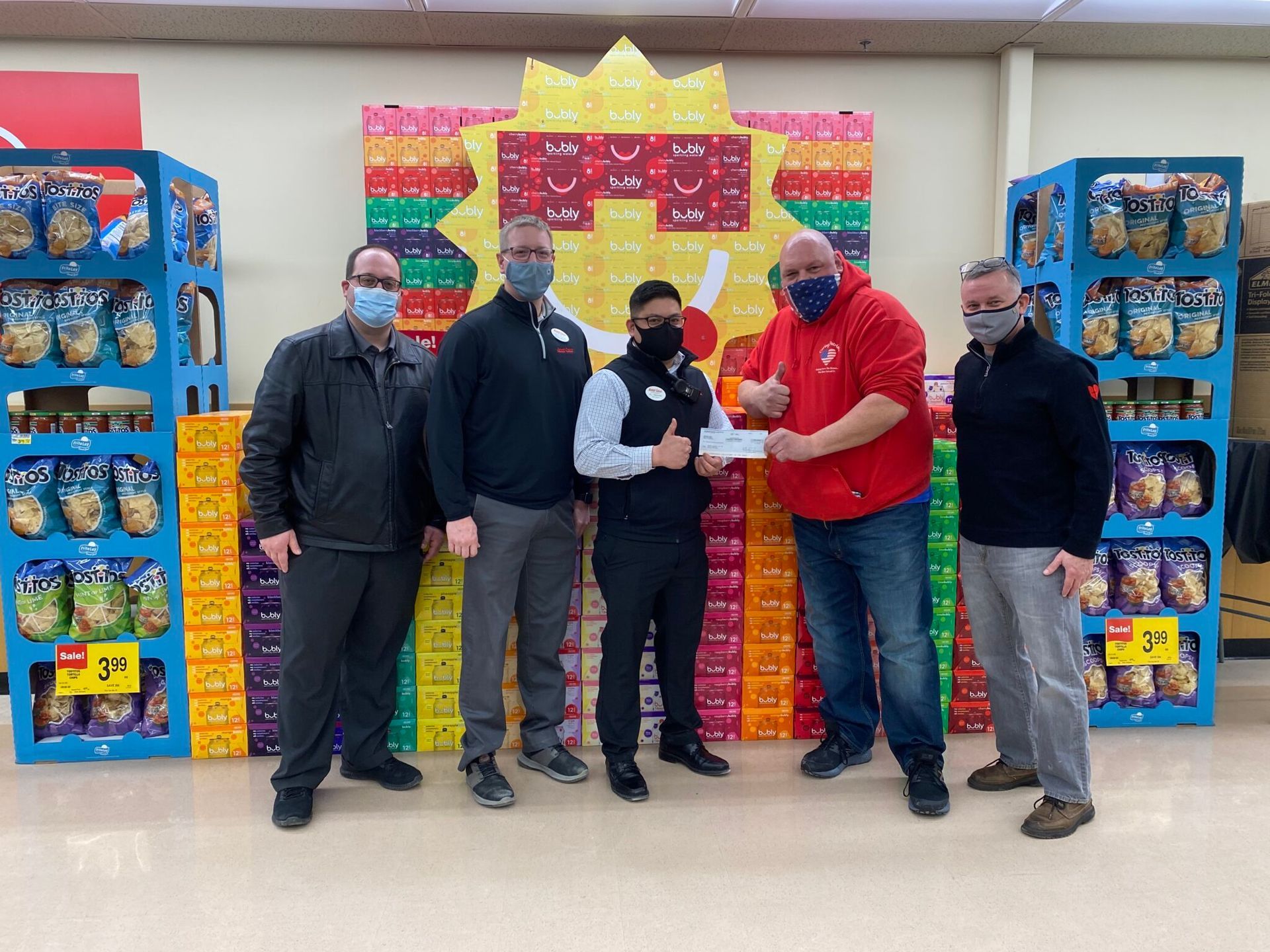 Four masked people posing in a store aisle beside stacks of colorful packaged goods and a gift display.