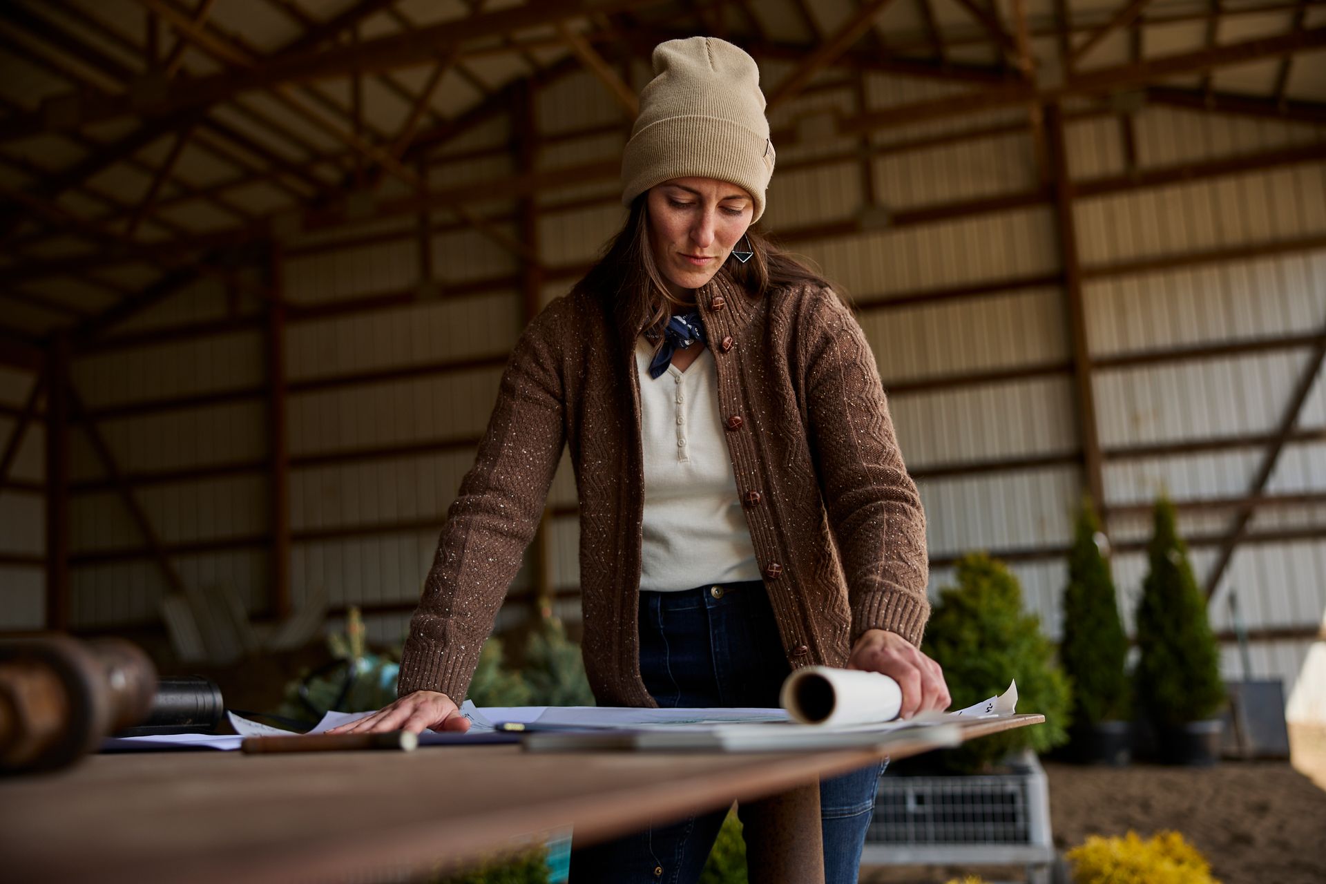 Person in a knit hat leans over a table reviewing a large blueprint in a workshop.