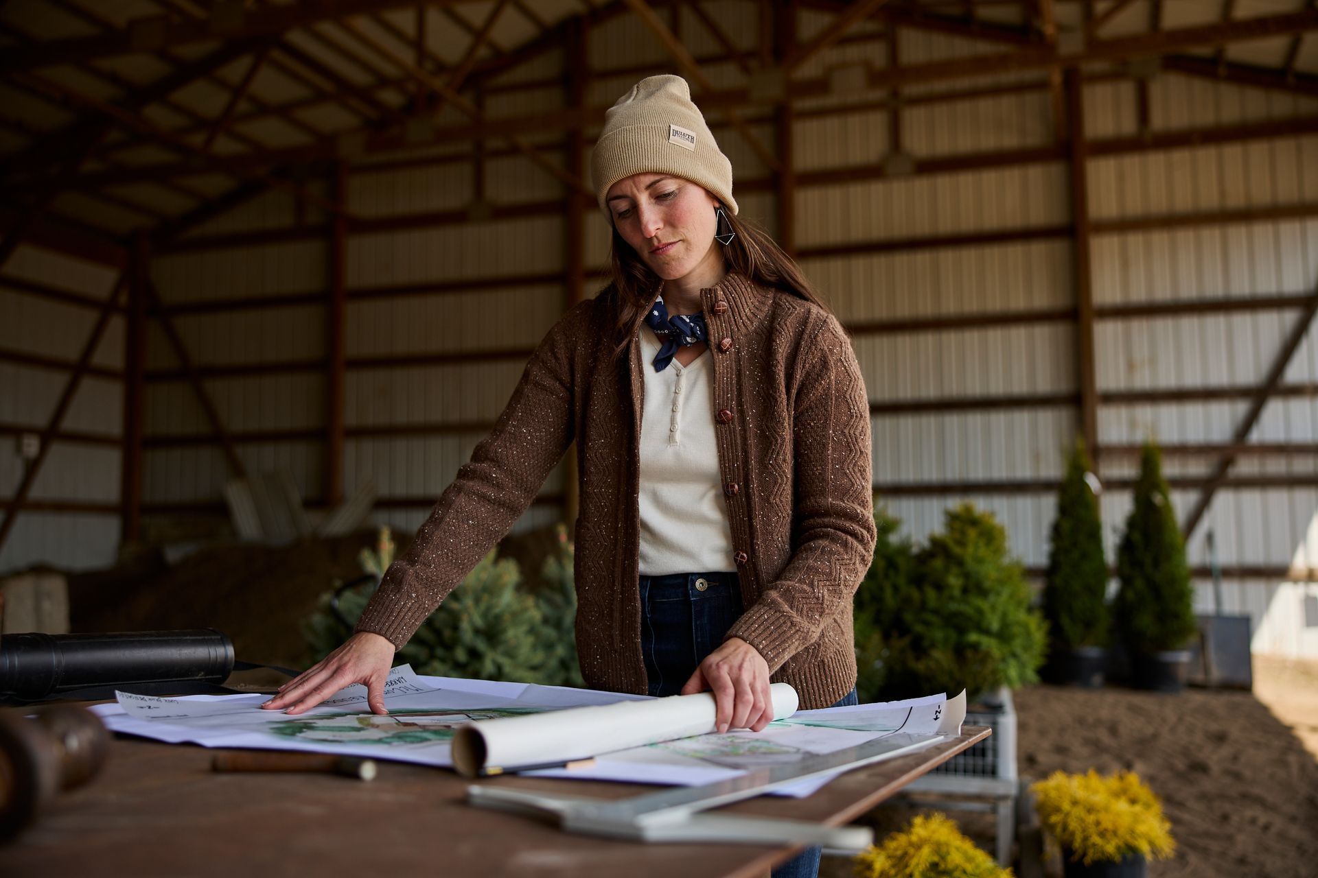 Person in a knit hat reviewing large architectural plans on a table in a barn-like workspace.