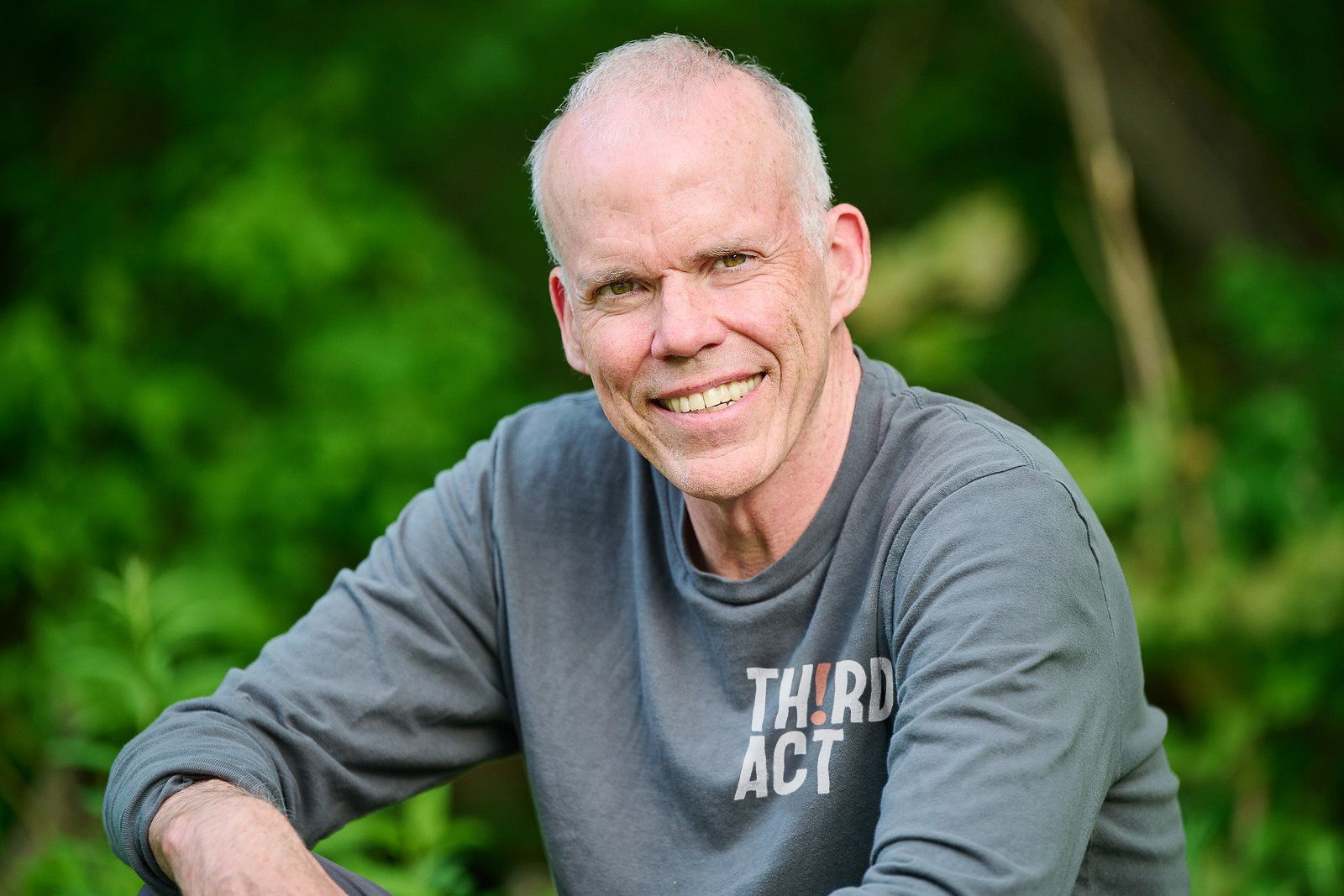 Smiling man in a gray shirt sitting outdoors against a green leafy background