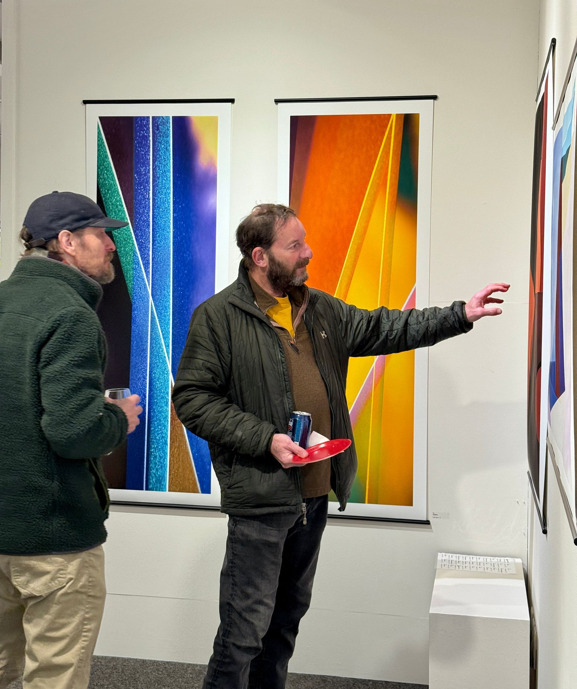 Two men in a gallery examine colorful vertical artworks on a white wall.
