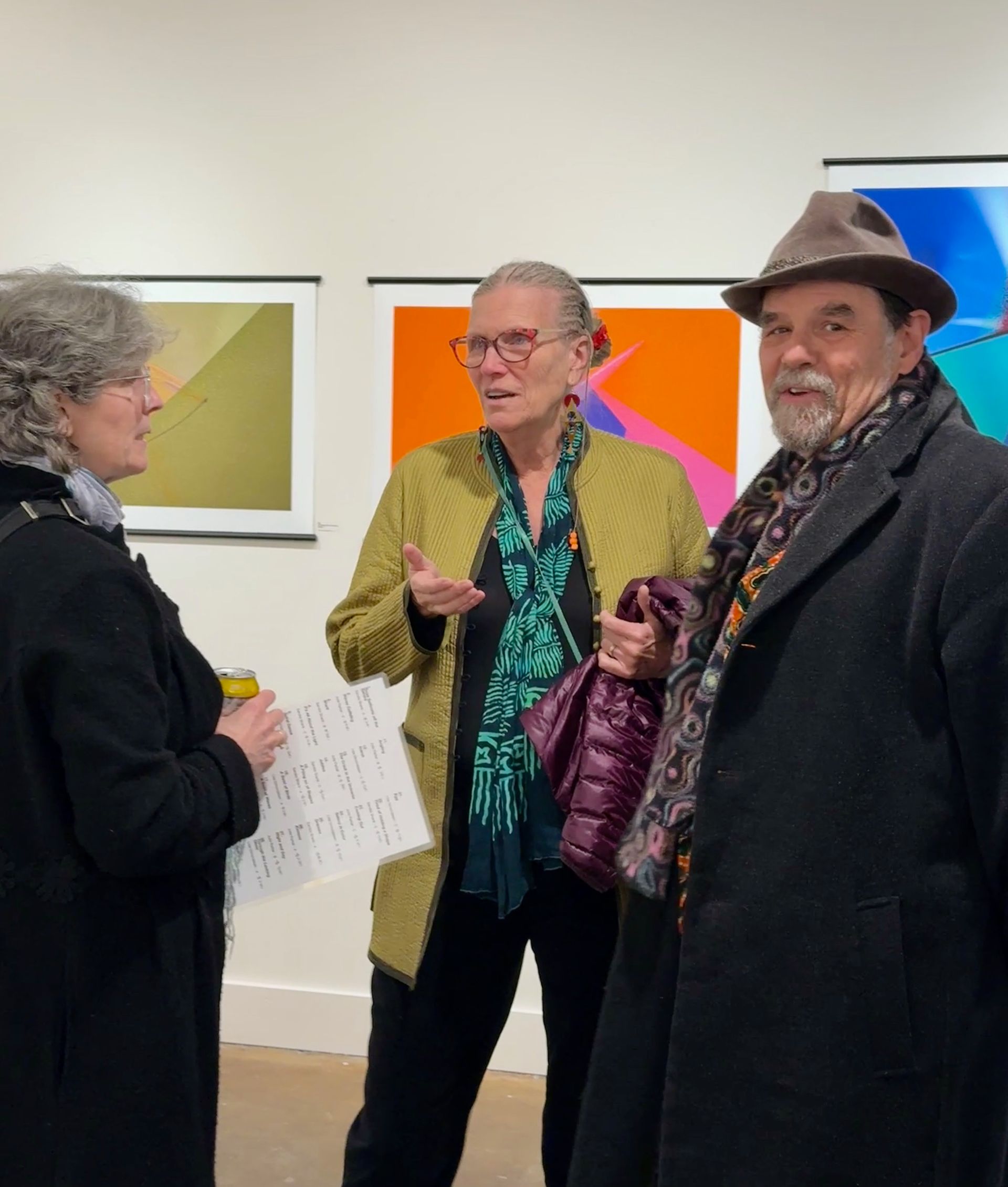 Three people chatting in an art gallery, with colorful framed artwork on the wall.