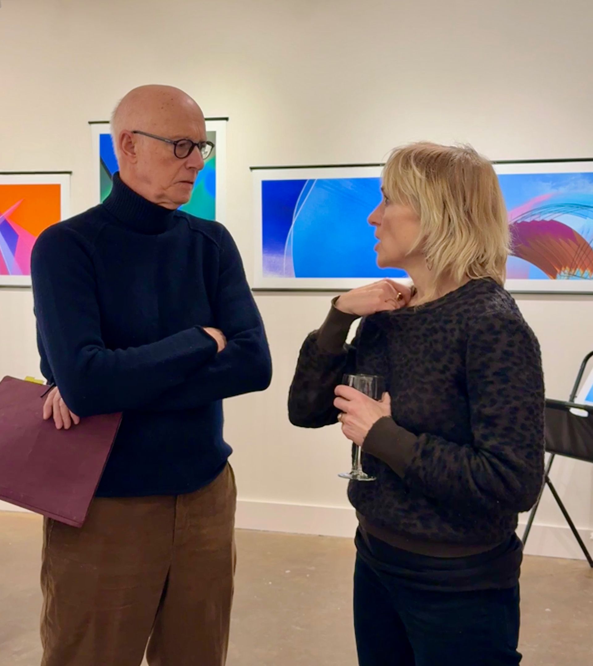 Two people talking in a gallery, with abstract colorful paintings on the wall.