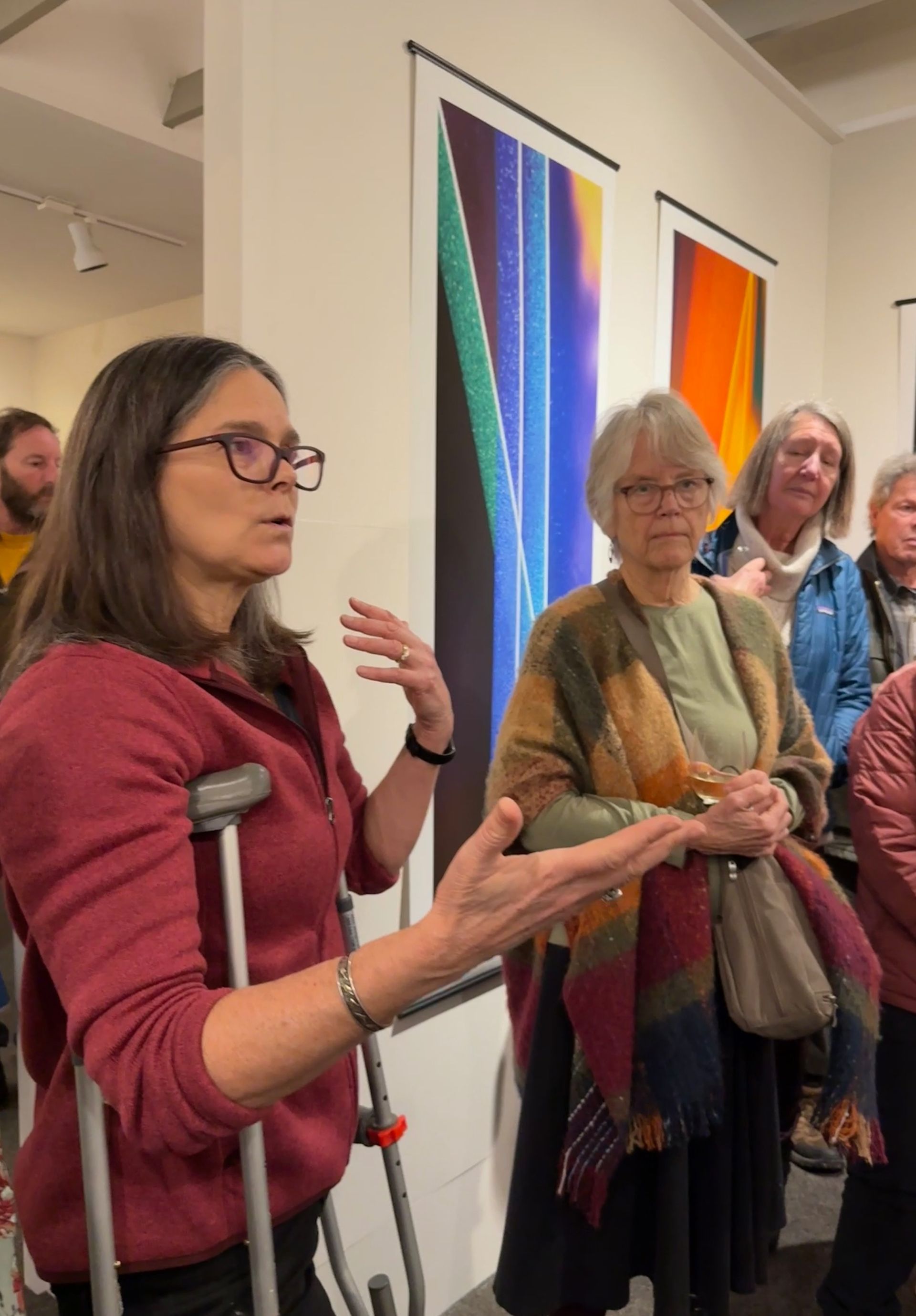Several people stand in a gallery beside colorful abstract paintings, listening to a woman on crutches speak.