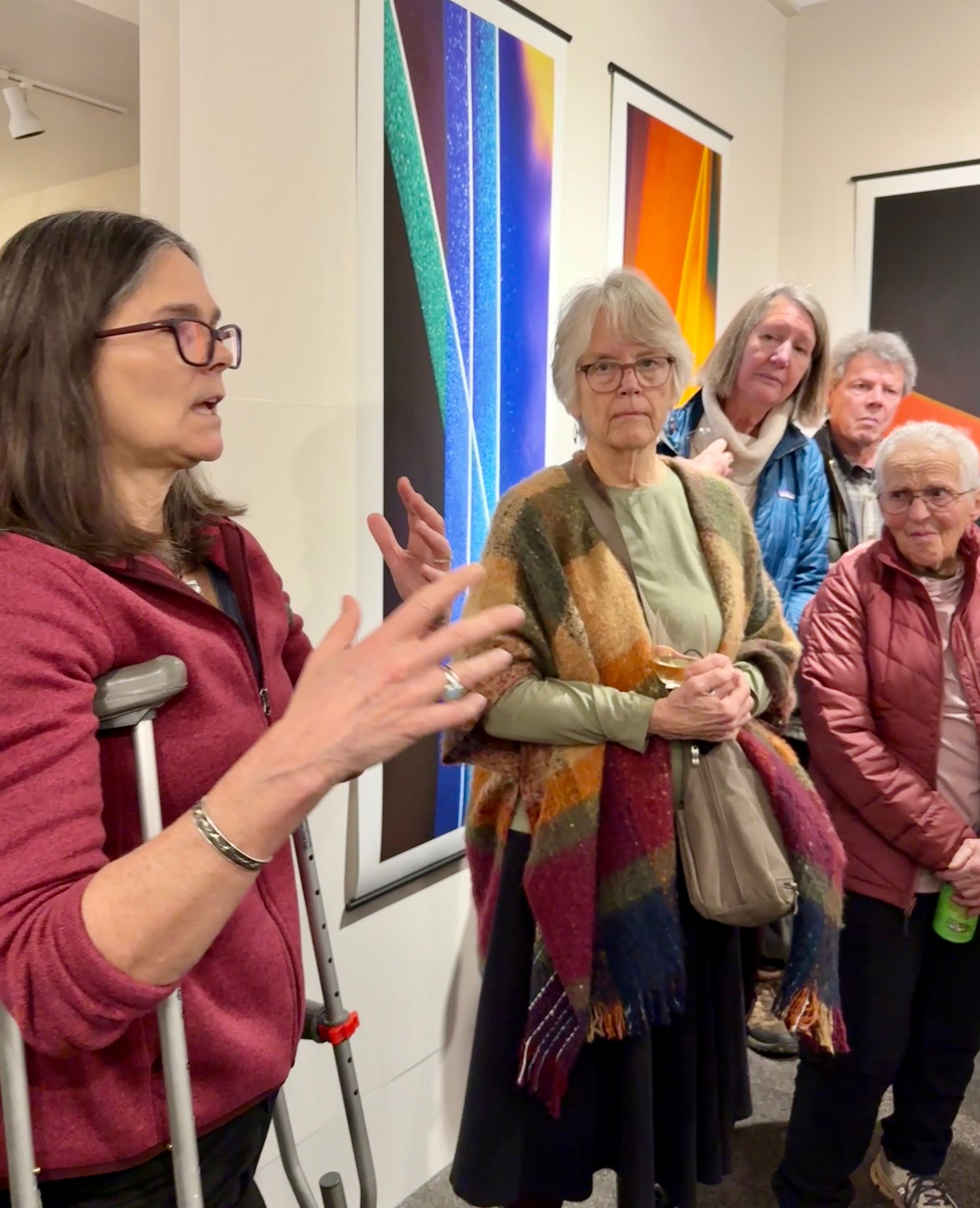 Woman speaking to a small group in an art gallery, with colorful vertical paintings on the wall.