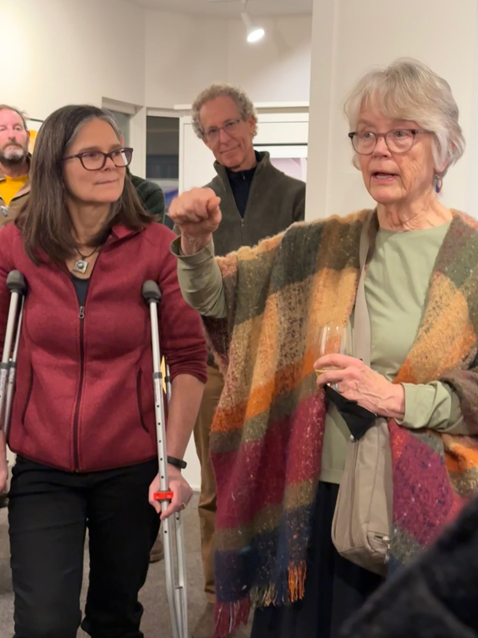 Two women with crutches talking indoors, one gesturing while others stand in the background.