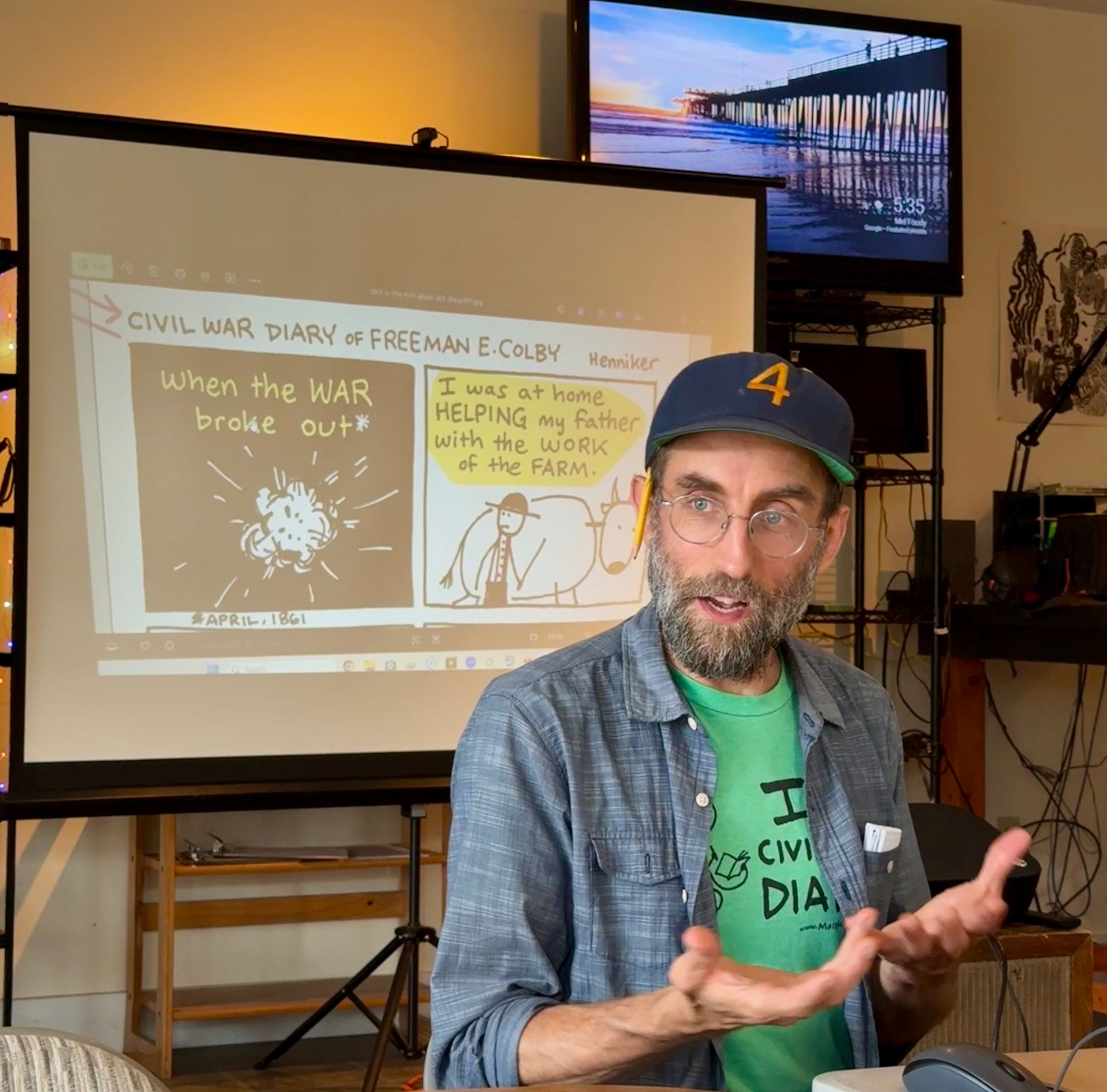 Presenter in a room beside a projected comic slide, gesturing while speaking.