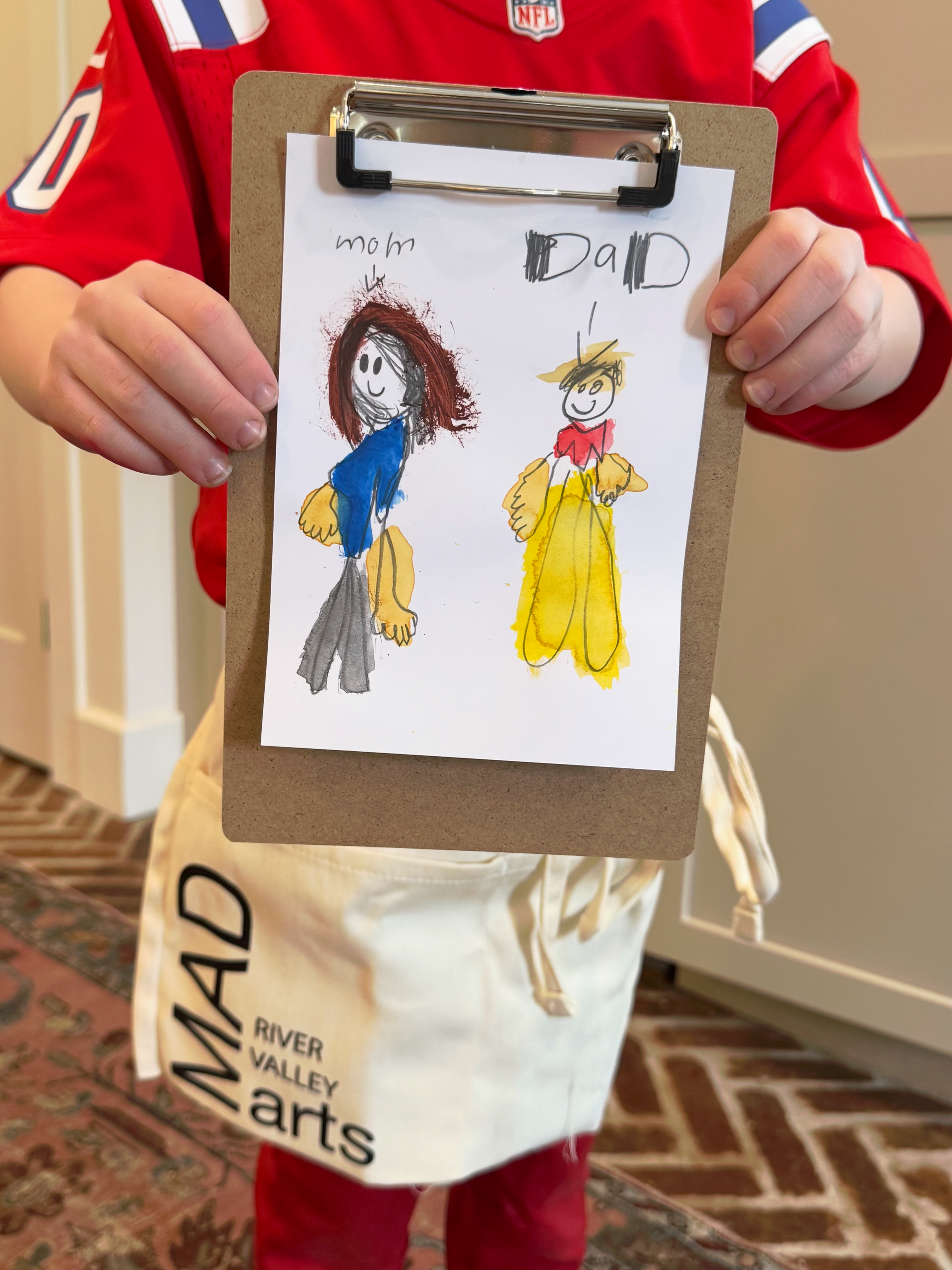 Child holding a clipboard with a crayon drawing in a beige indoor hallway, wearing a red shirt and art apron