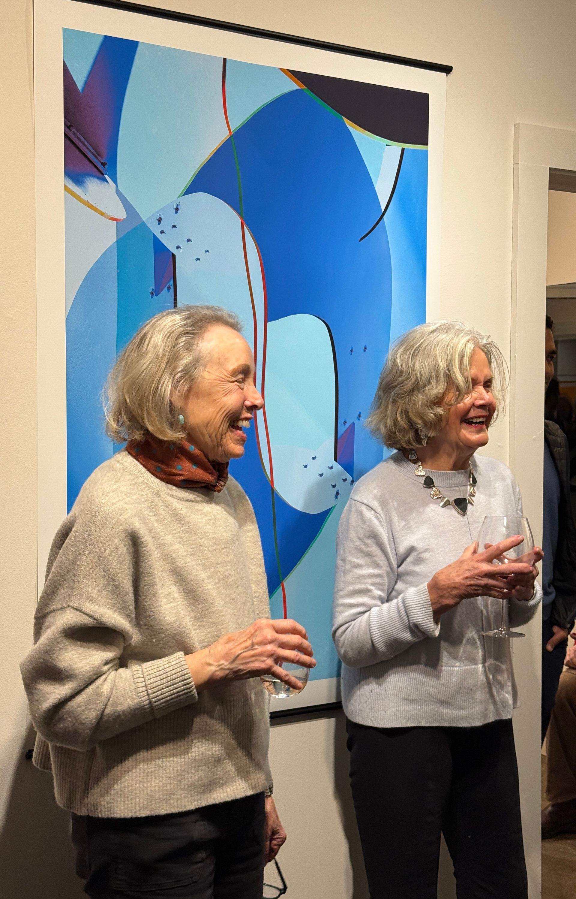 Two smiling people standing indoors beside a large blue abstract painting