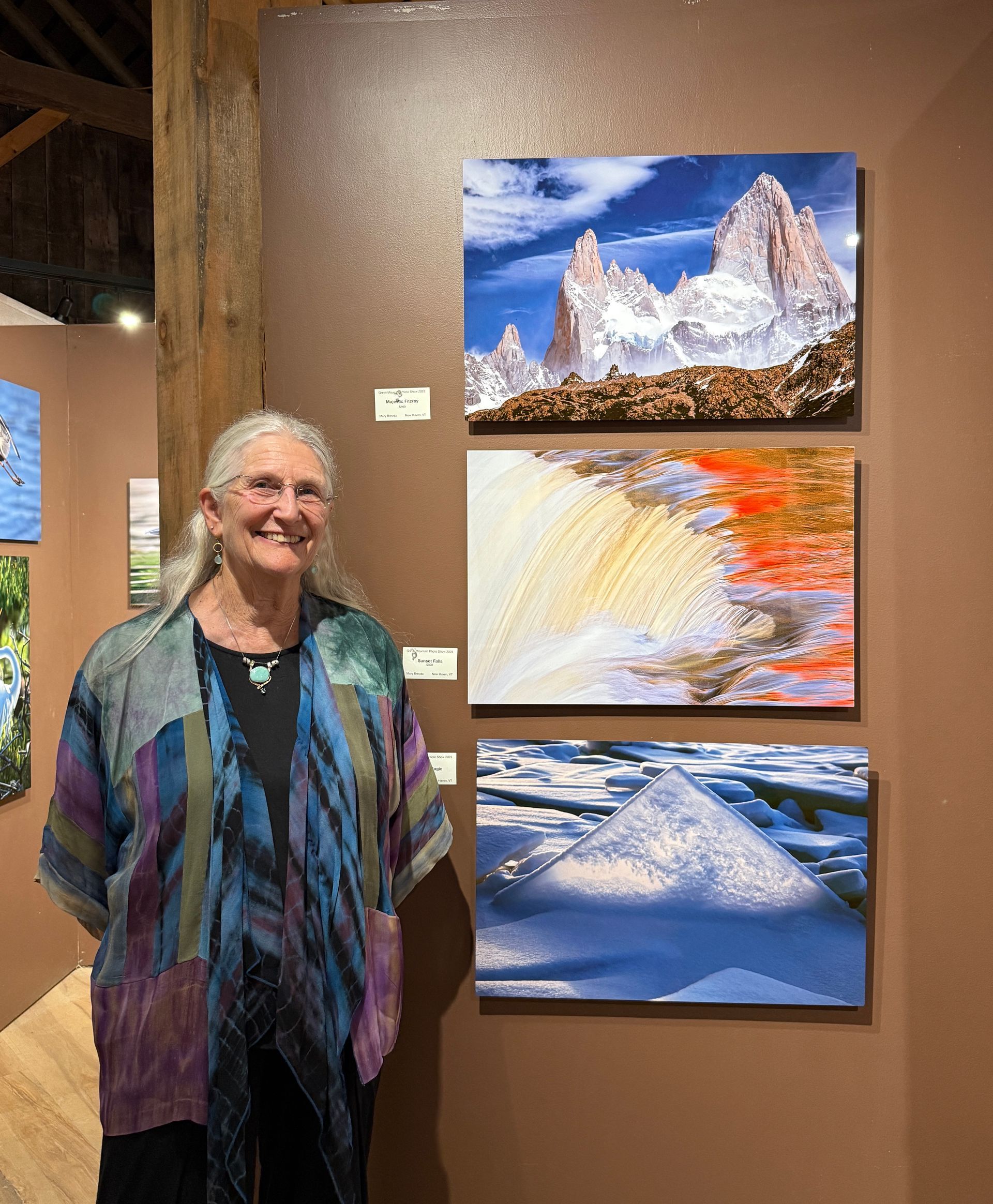 Smiling woman stands beside three colorful landscape paintings in an art gallery.