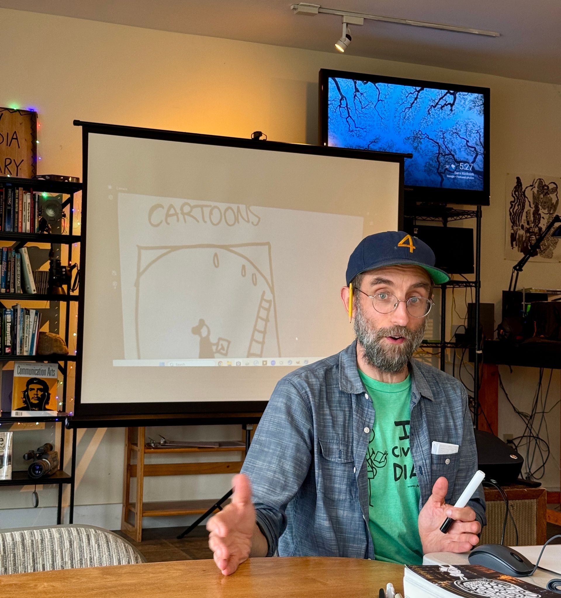 Man speaking at a table in a room with a projected cartoon sketch on a screen behind him.