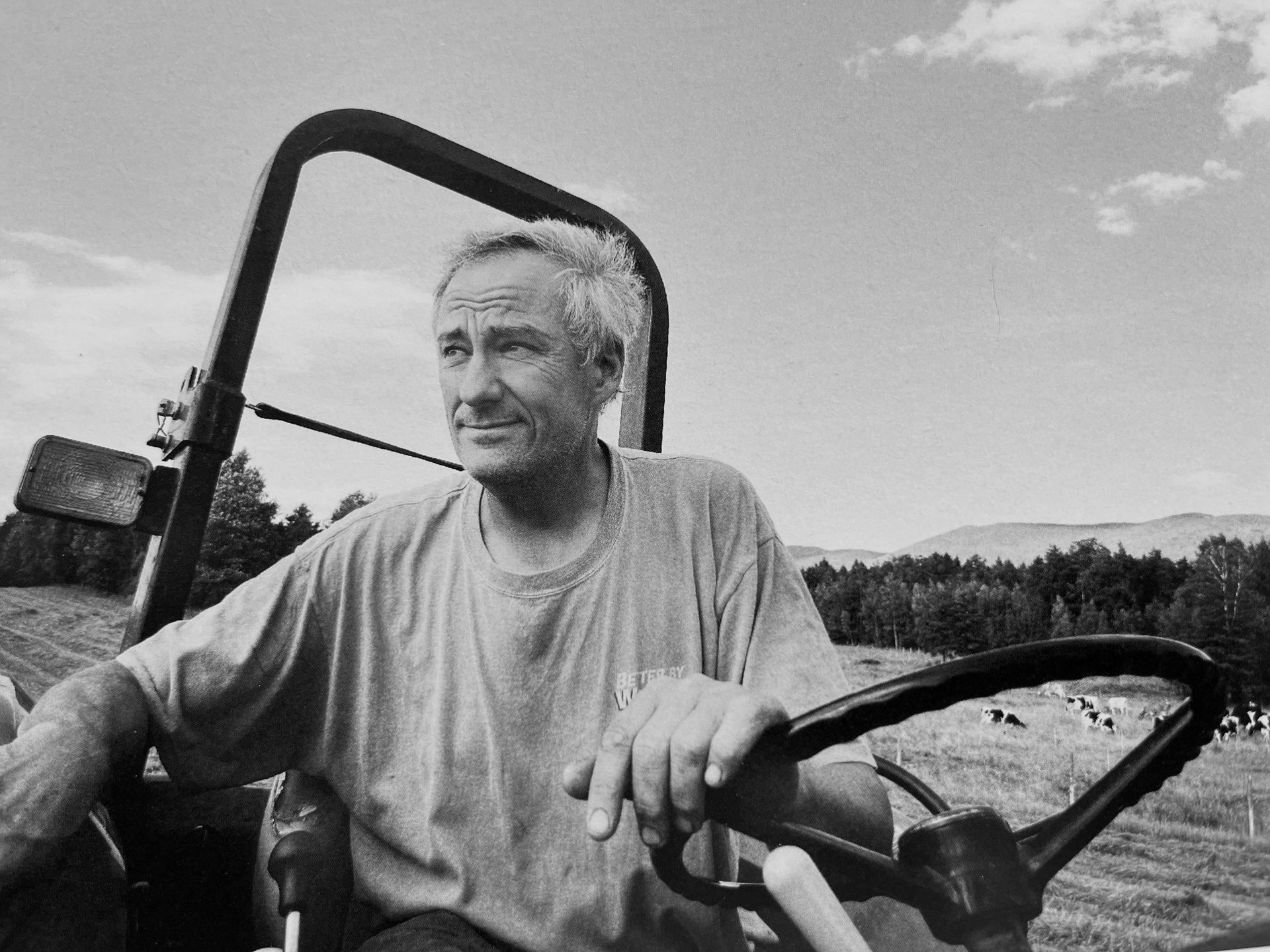 Man seated in an open vehicle, smiling beside a steering wheel in a rural field under a cloudy sky.