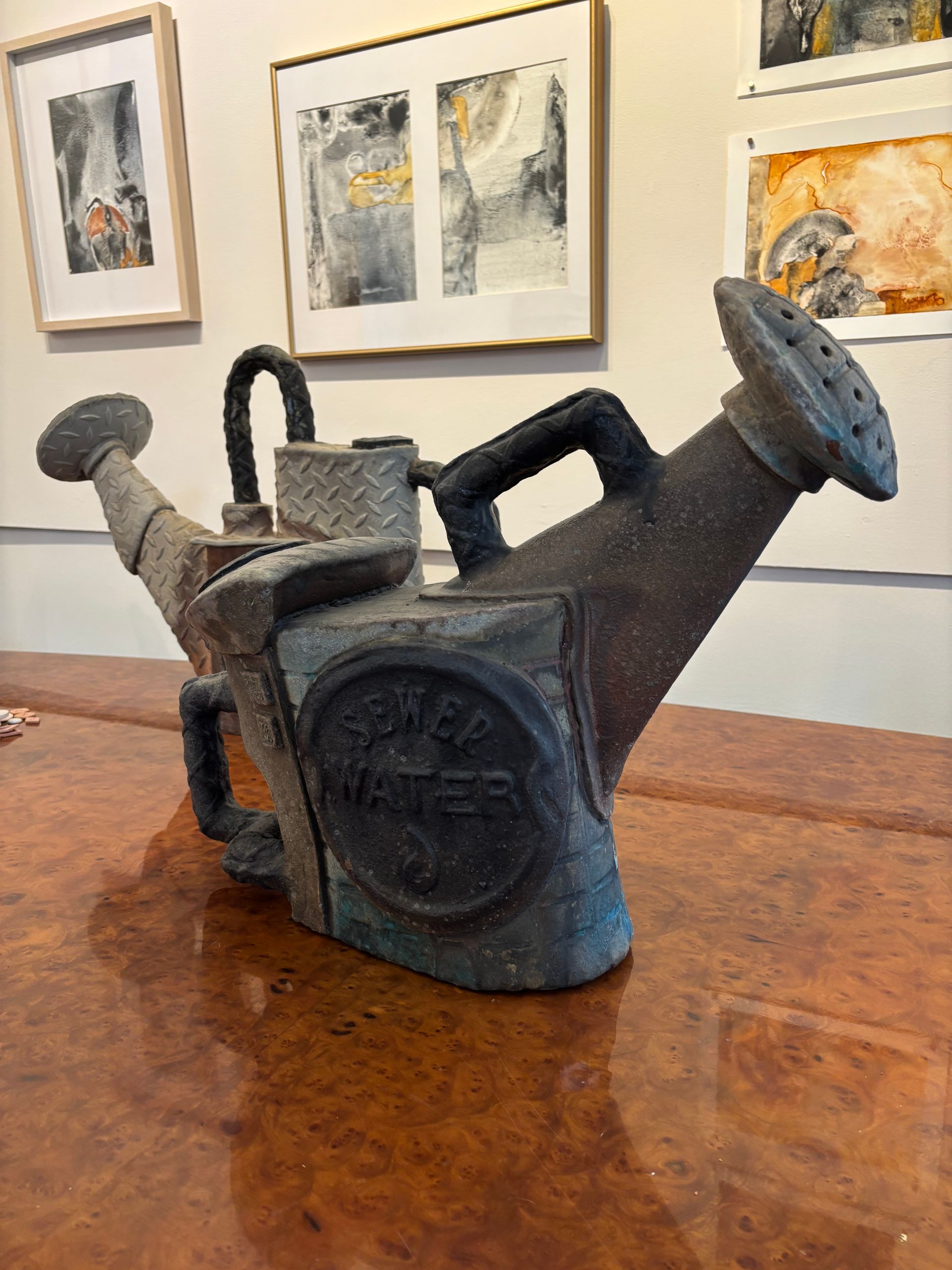 Antique cast-iron watering can sculpture on a wooden table in a gallery, with framed artwork in the background