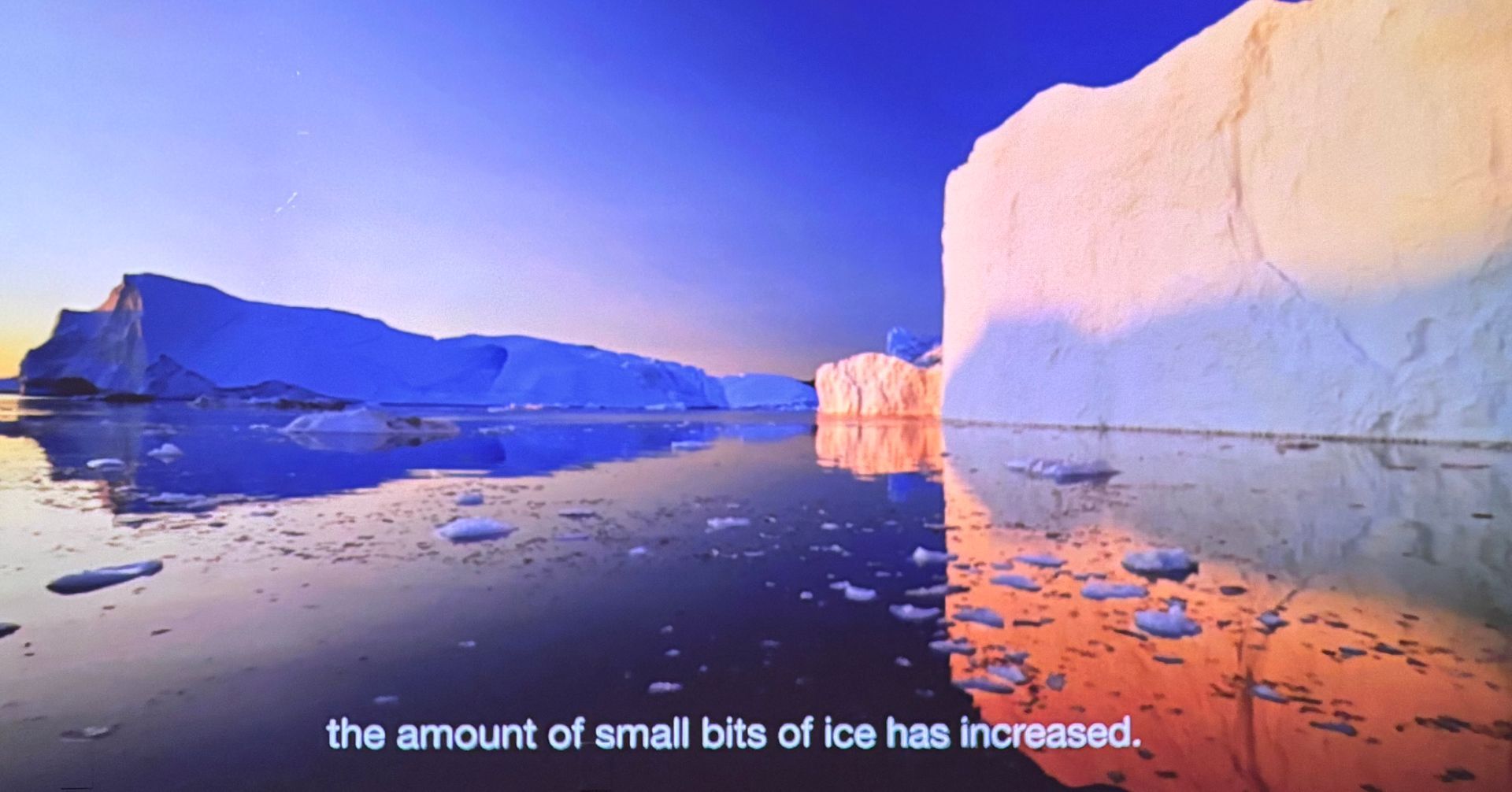 Icebergs and glacier cliffs reflected in calm water at sunset, with small ice pieces in the foreground.