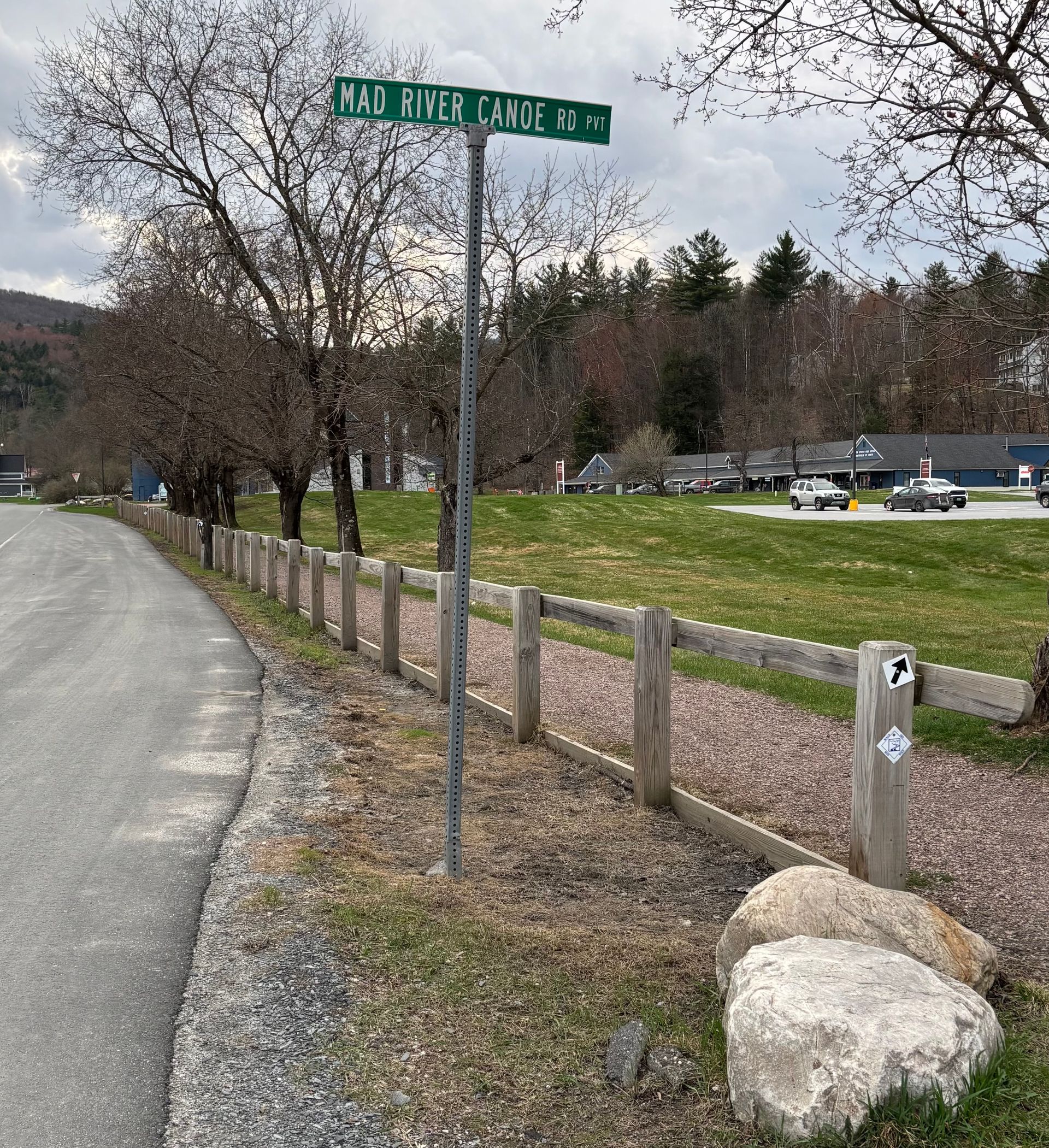 Rural trail entrance with wooden fence, gravel path, signpost, grassy field, and parked cars under cloudy sky