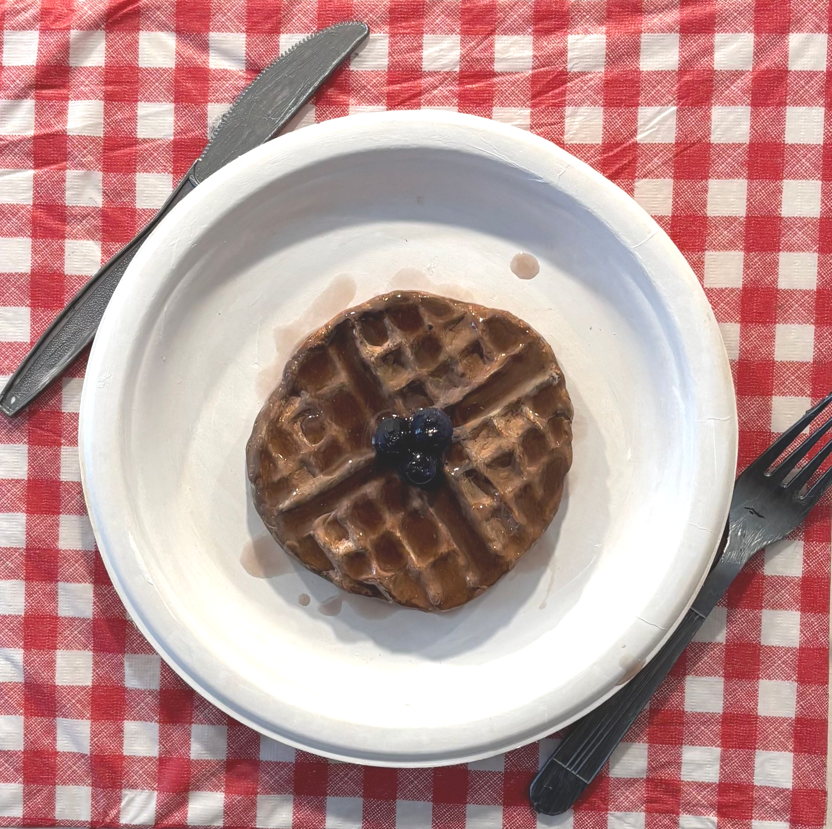 Waffle on a white plate with a dollop of dark berry jam, on a red-checkered tablecloth with fork and knife nearby