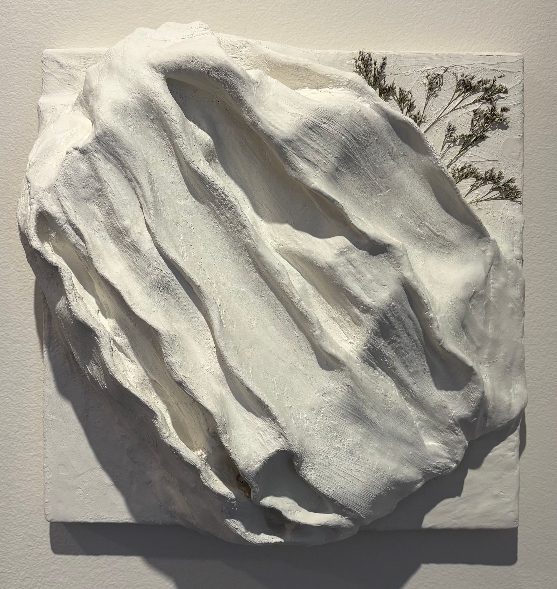 Abstract white textured wall sculpture on a square panel with small tree branches in the top right.