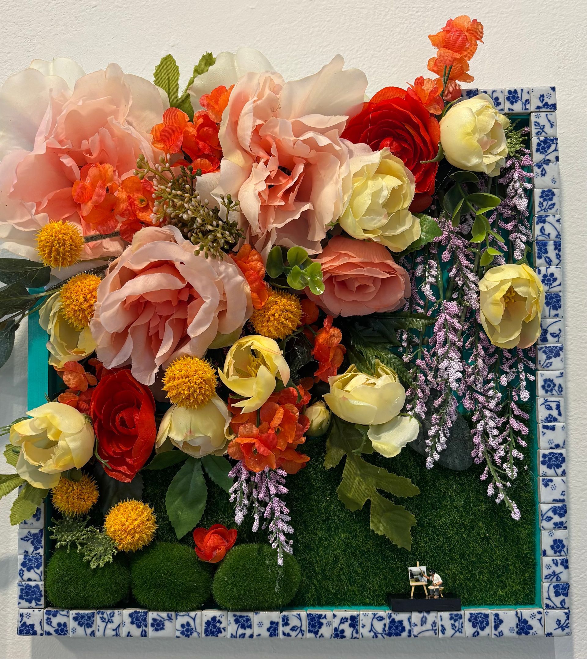 Floral wall arrangement with pink, red, yellow, and white flowers on a green background, framed in blue tile.