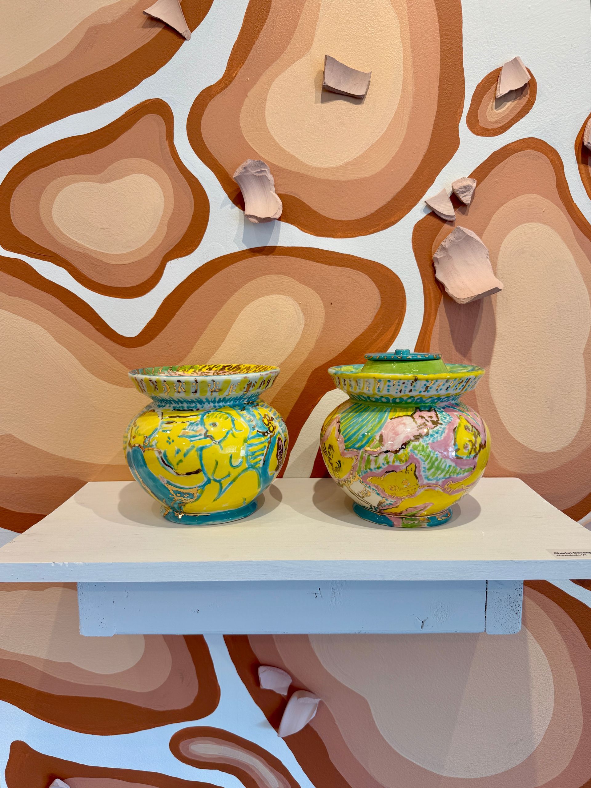 Two colorful patterned jars on a white shelf against an abstract tan-and-brown wall.