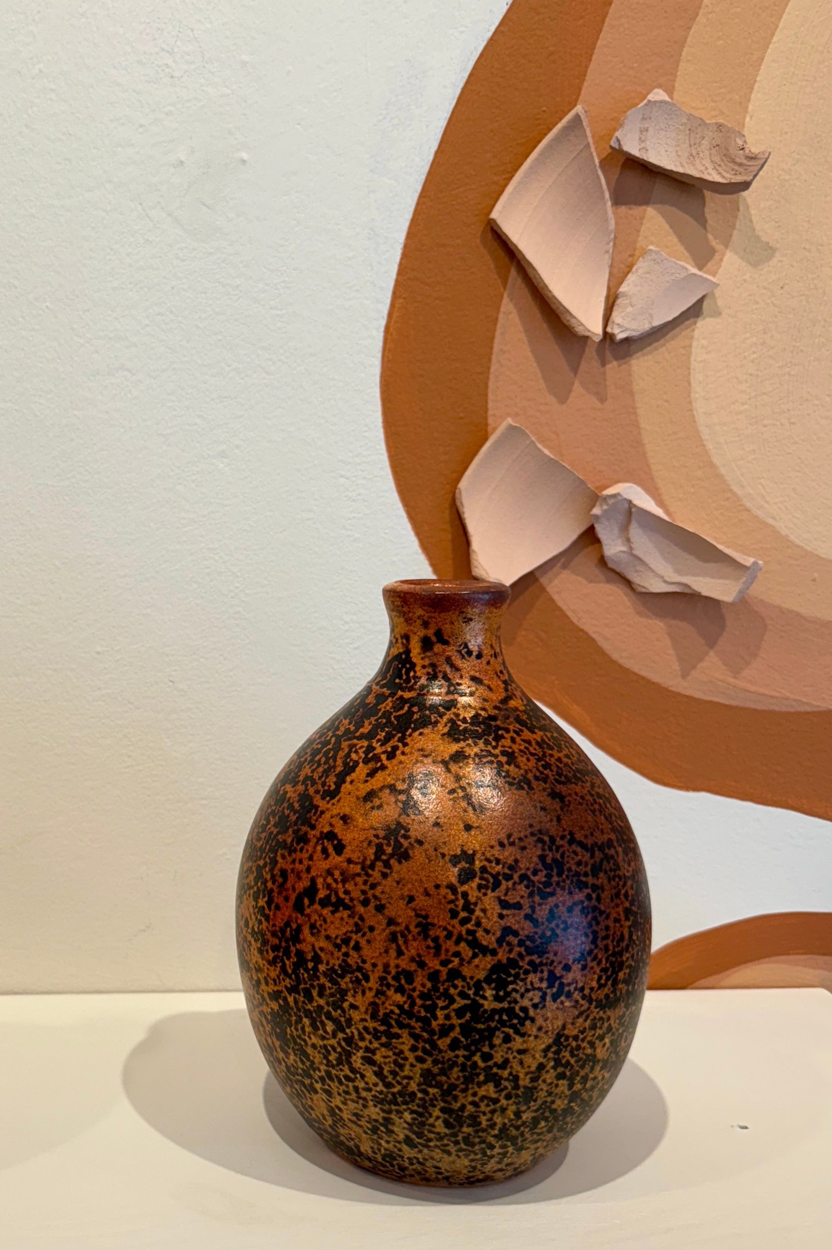 Speckled brown vase on a white surface in front of a beige abstract wall decoration