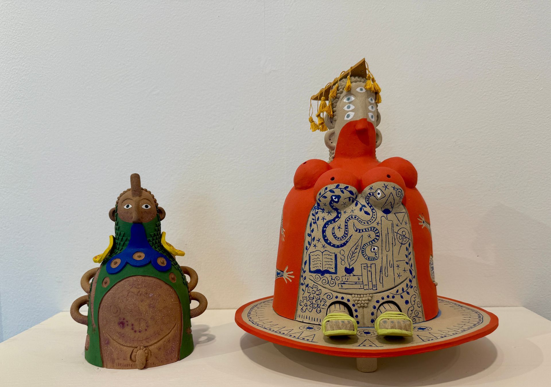 Two decorated figurines on a white surface: a small brown-blue figure and a larger orange-white figure on a plate.