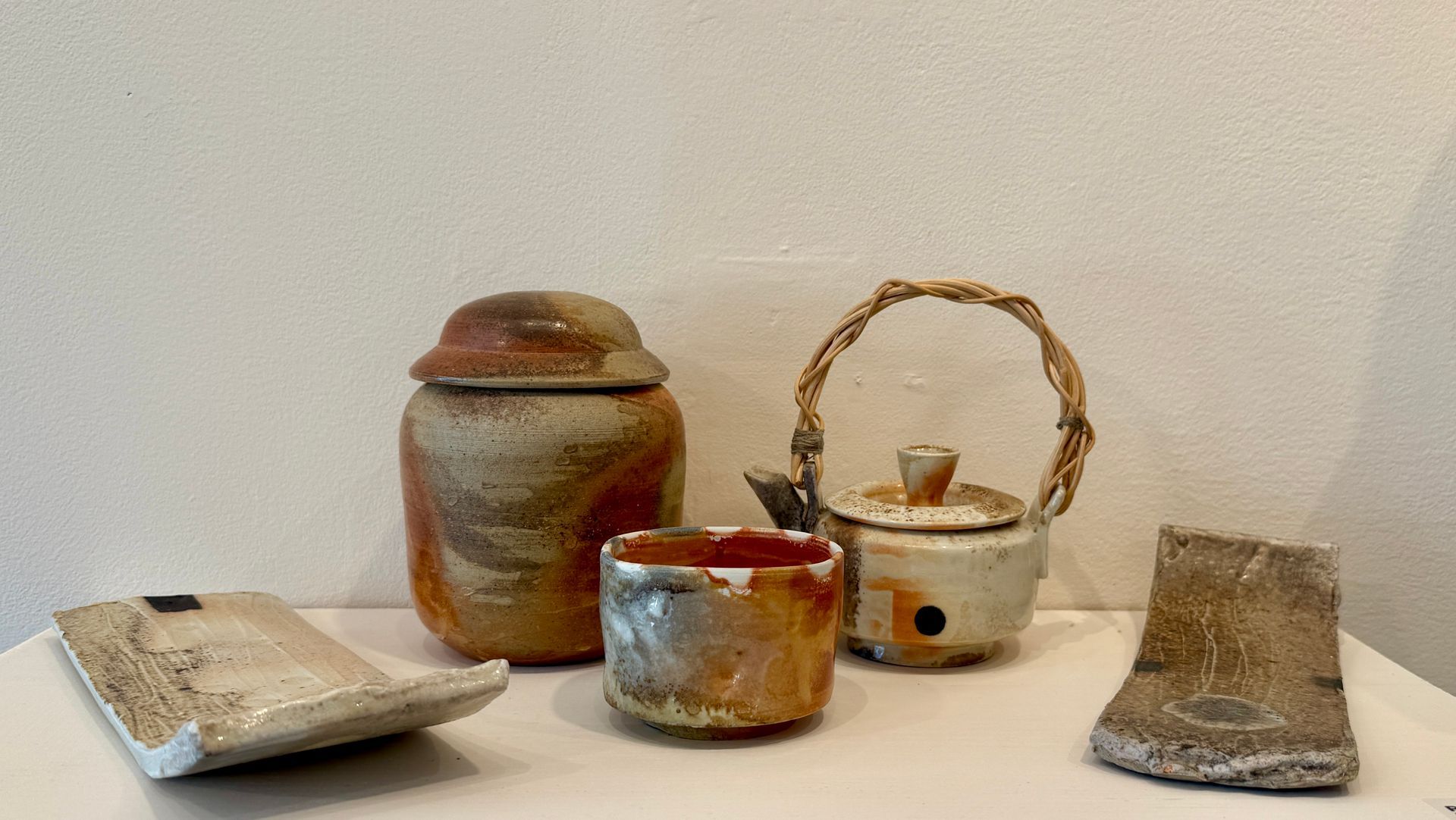Ceramic jars, cups, and a woven basket on a white surface against a plain wall