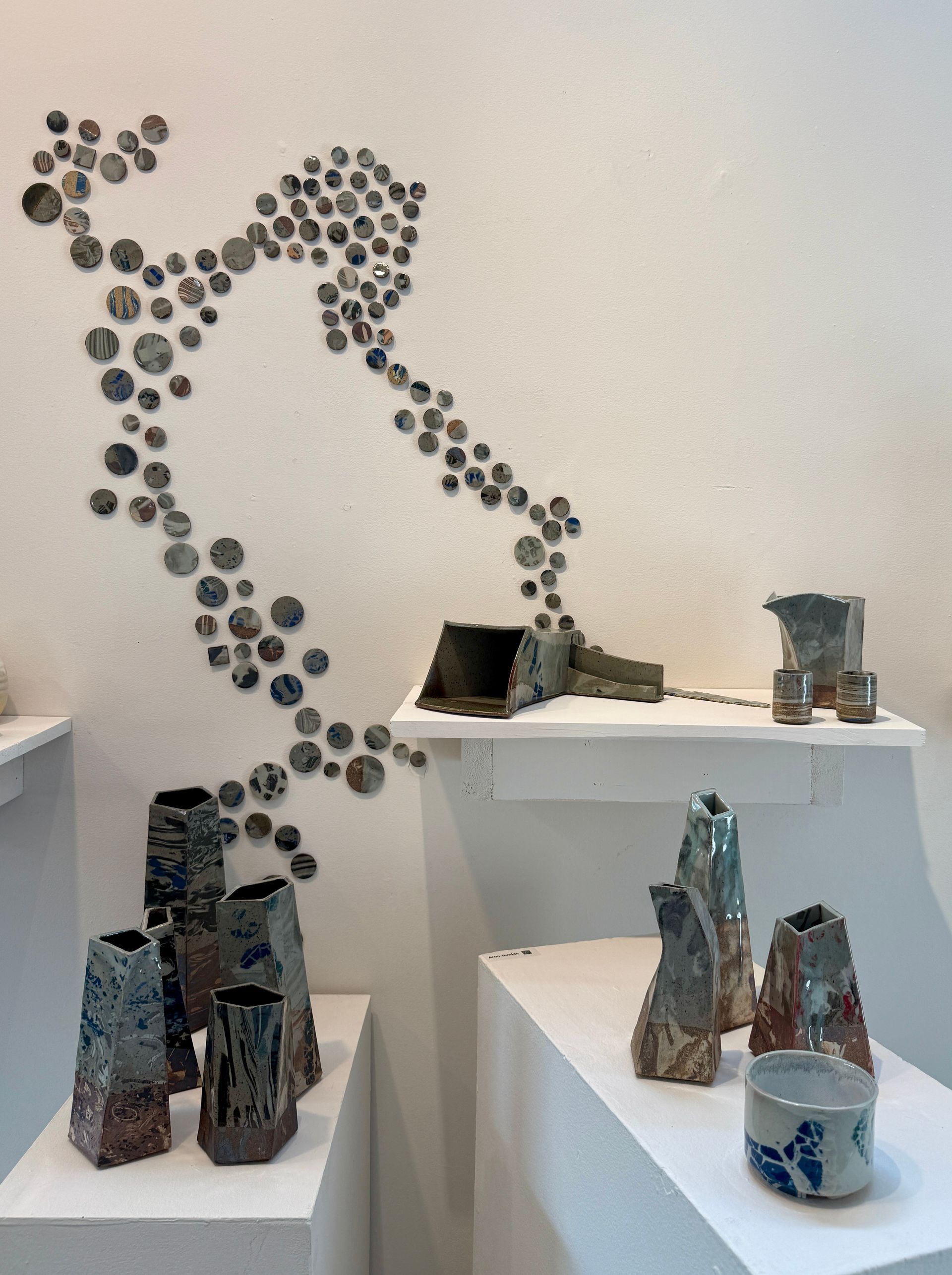 Art gallery display of ceramic sculptures on white pedestals with a dot-pattern wall installation behind them