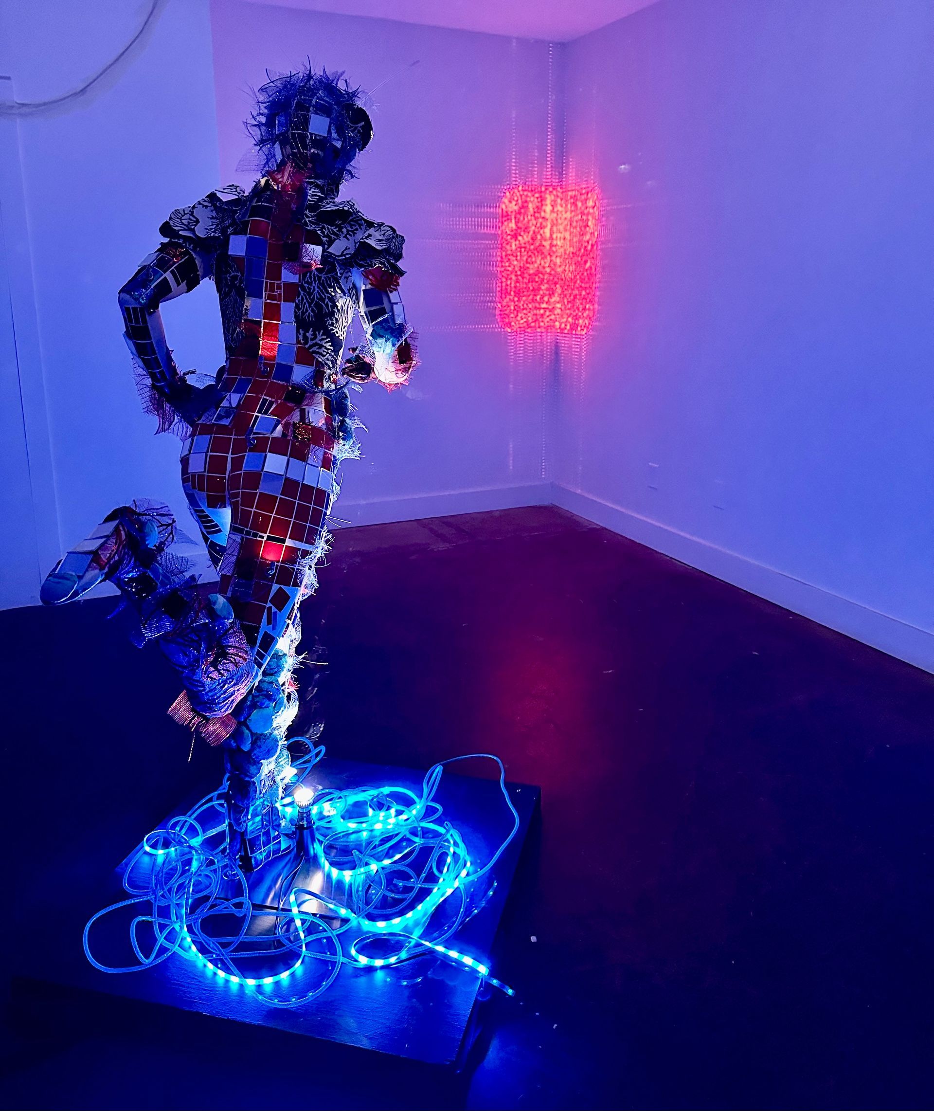 Colorful illuminated sculpture of a human figure standing in a dark room, with blue lights at the base and red lights on the wall