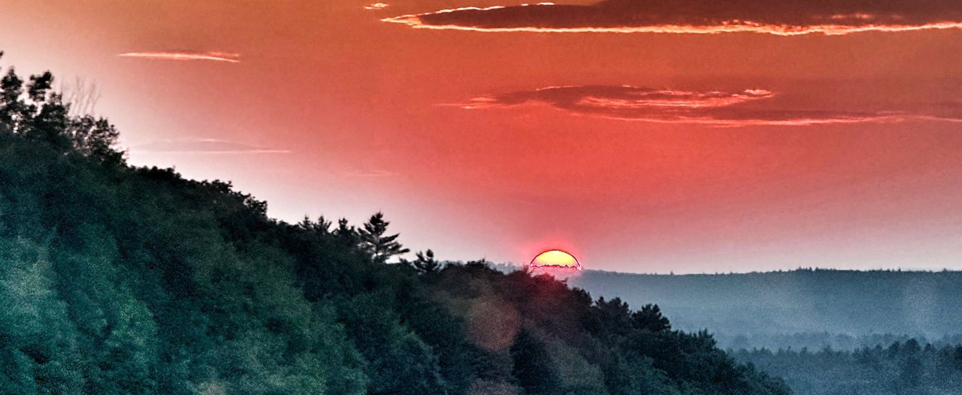 Sunset over forested hills with a red sky and the sun dipping below the horizon