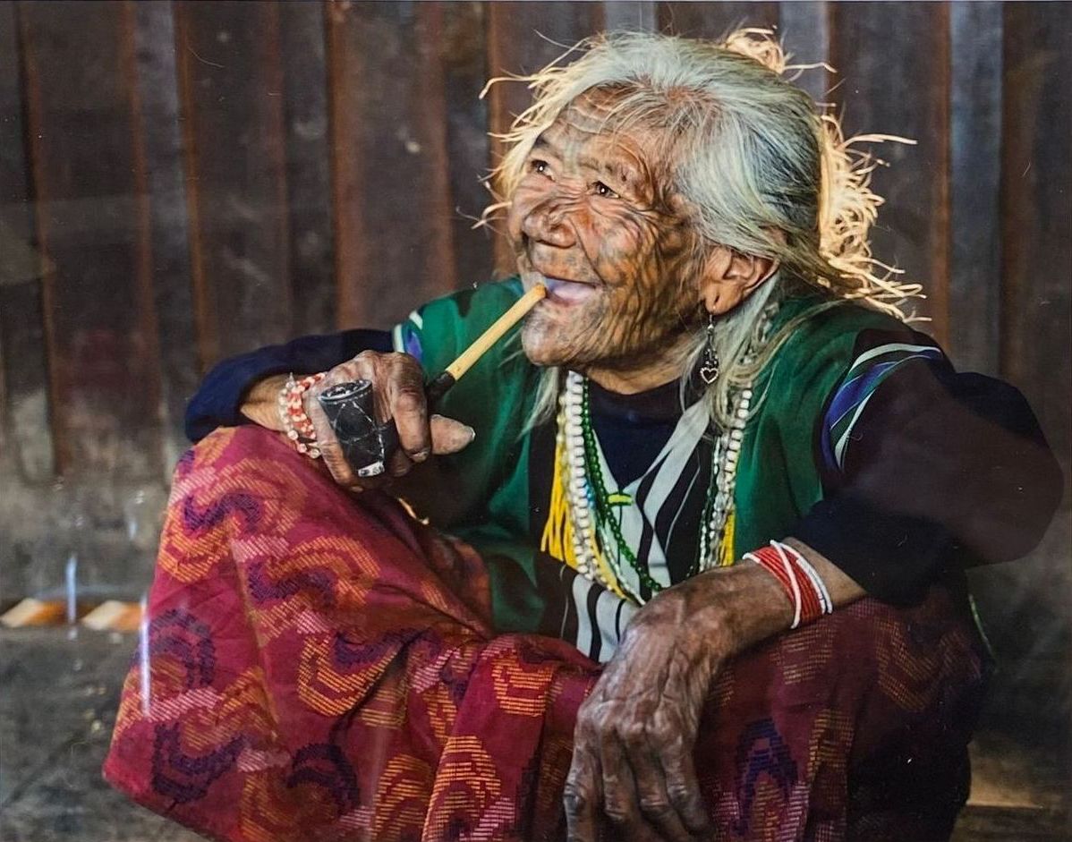 Smiling person with white hair sits cross-legged, holding a cigarette, in colorful layered clothing.