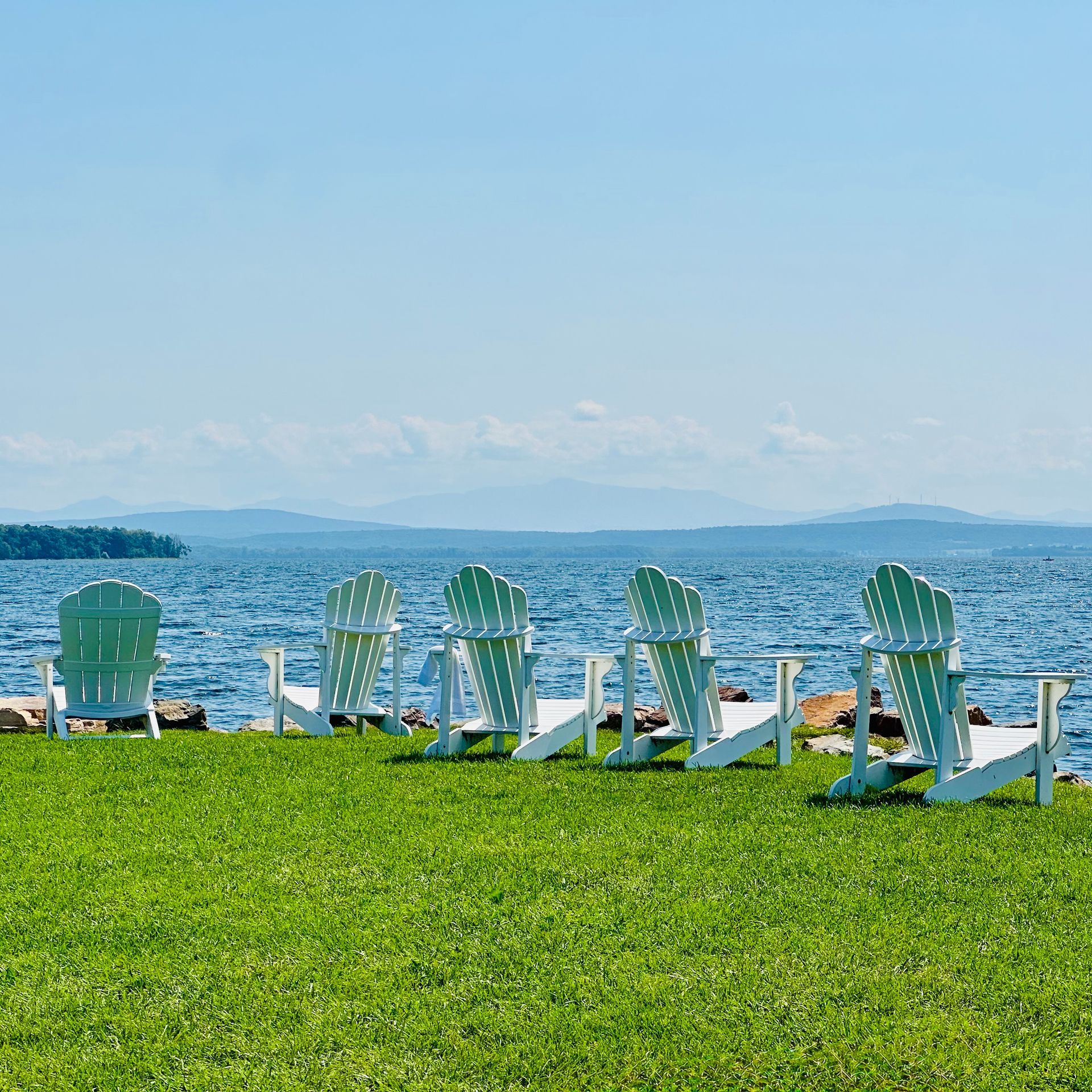 Empty green lawn with white Adirondack chairs facing a blue lake and distant mountains under a clear sky