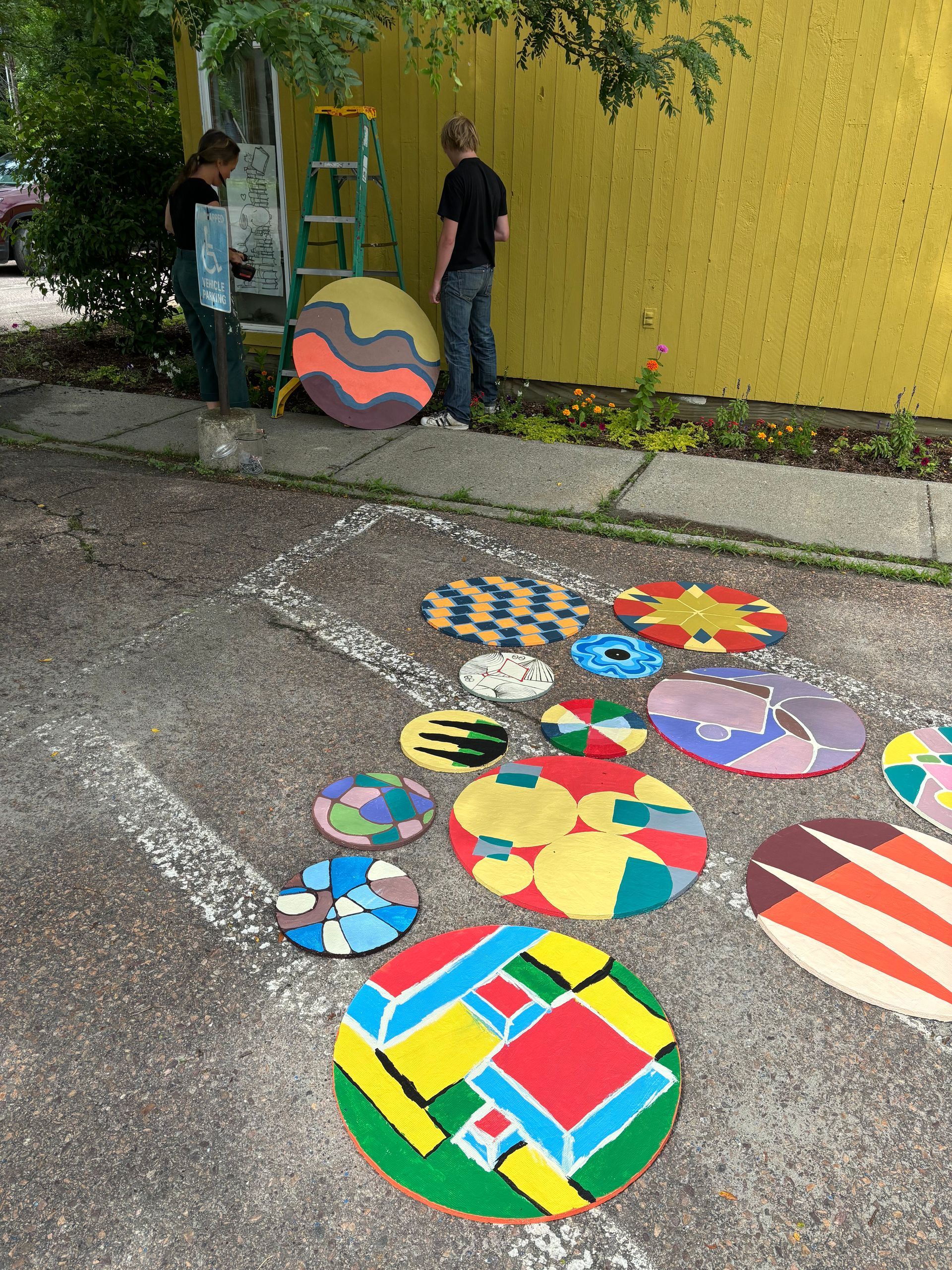Person stands beside colorful painted stones outside a yellow building, with more painted stones arranged on the ground.