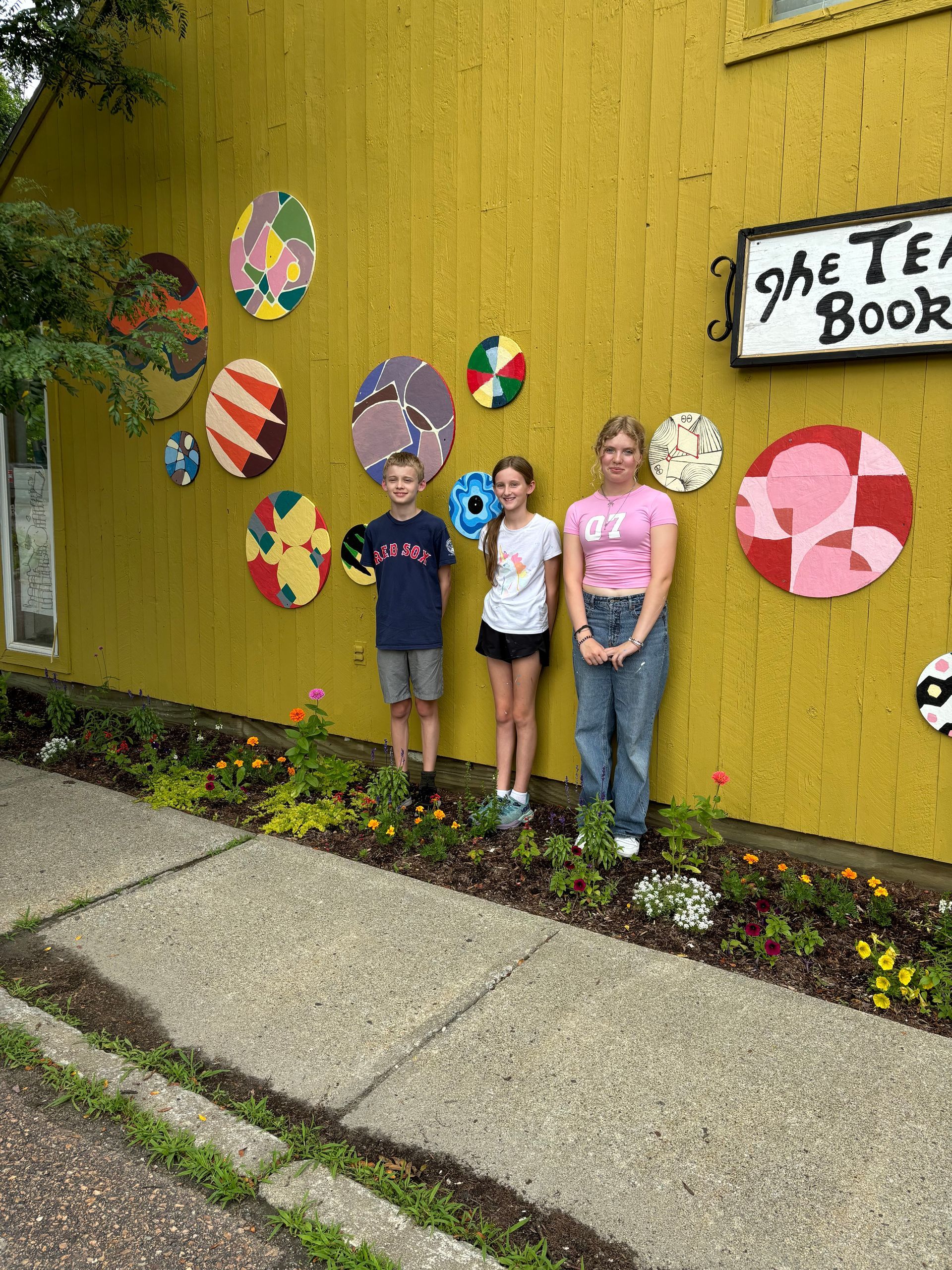 Three children stand by a yellow wall with colorful painted circles and a sign reading “The Tea Book.”