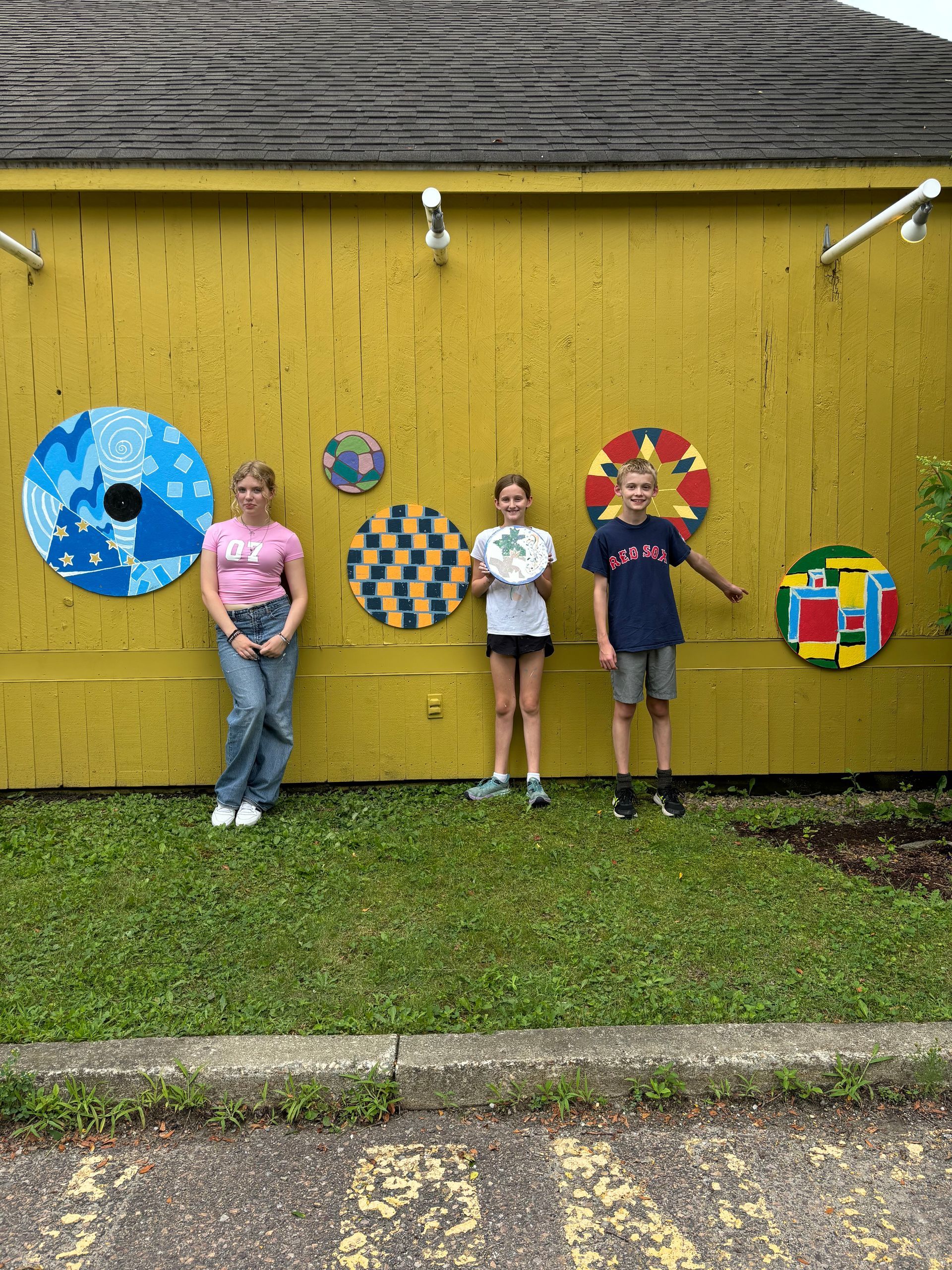 Three children standing by colorful circular art on a yellow wall outdoors
