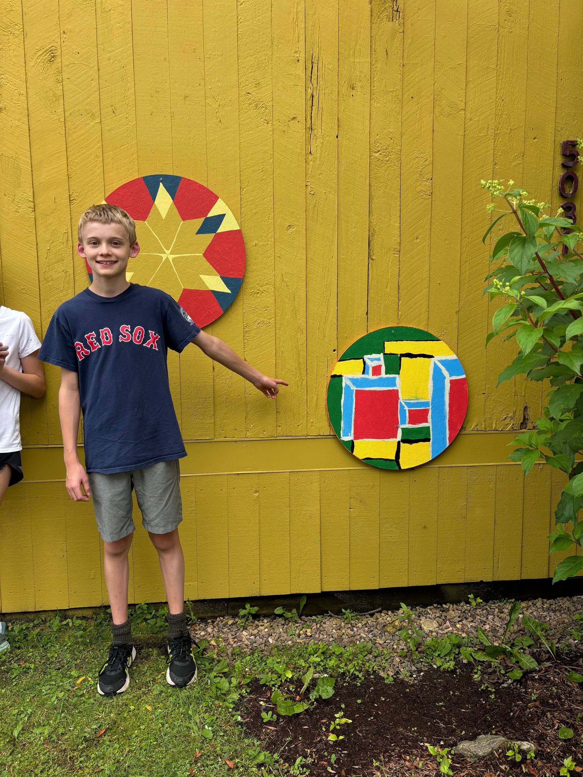 Child standing by yellow wall with colorful circular mosaics and greenery nearby